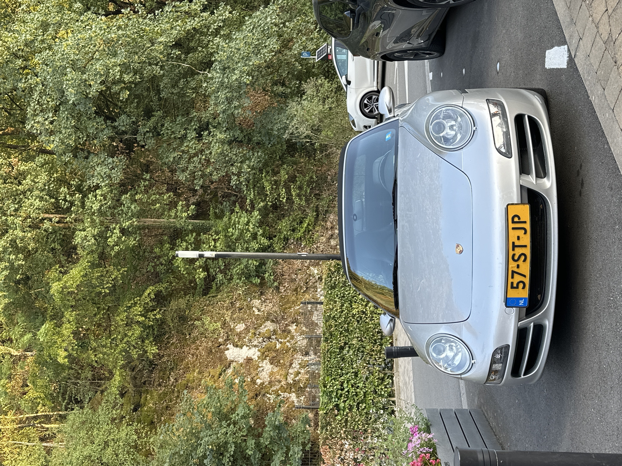 The image depicts a silver Porsche 911 parked on an asphalt surface, likely a street or parking area, in Durbuy, Belgium. The car's license plate reads "NL 57-ST-JP". To the right of the Porsche, a dark-colored SUV is partially visible, also parked. In the background, a white car is parked further down the street, and a blue parking sign with a white "P" is visible. The environment is characterized by a lush, green hillside densely covered with trees and foliage, with a rocky outcrop visible in places. A neatly trimmed green hedge runs along the base of the hillside, separating it from the paved area. In the foreground, a planter with pink and white flowers is situated to the left of the Porsche. The lighting suggests it is daytime, with sunlight illuminating the scene and creating shadows. The scene is static, with no visible people or activity.