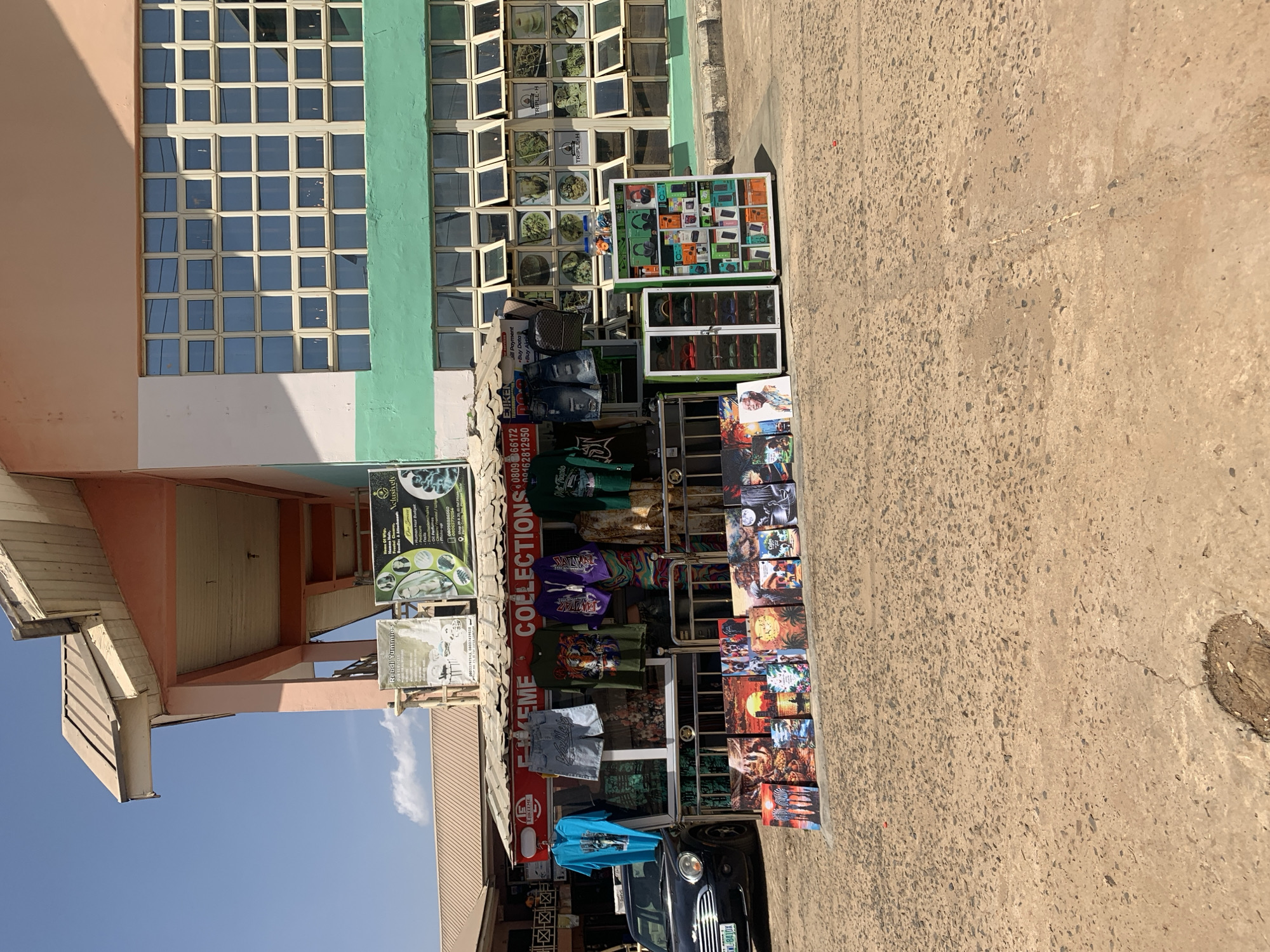 The image shows a storefront in Rafin Par, Nigeria, offering a variety of goods and services. The building has a facade with peach, green, and white paint. A large window with many panes is visible. The storefront displays clothing items, including T-shirts and shorts, along with other merchandise. Signs advertise "Extreme Collections", "Rebdi Yummies", "Nelusively Make-up", and "Triple H". Phone numbers are prominently displayed on a sign: "080966172" and "00162812950". A black car is parked in front of the store. The street is paved with concrete.