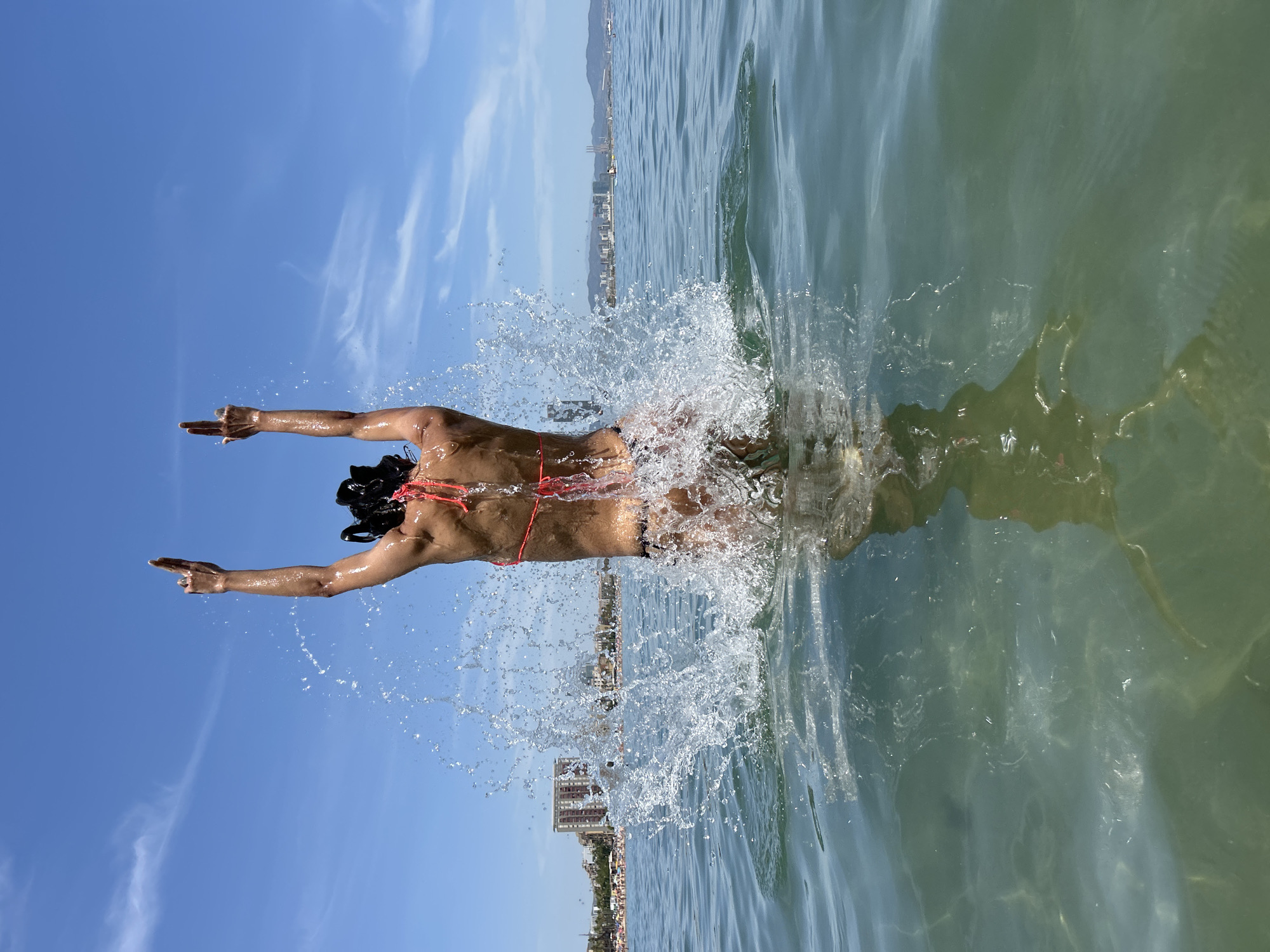 In Barcelona, Spain, a person in a bikini jumps into the water creating a large splash. The person's back is visible, with arms raised above their head. Buildings and a beach can be seen in the background on a sunny day with a blue sky and white clouds. The water is clear with sunlight reflecting on the surface.
