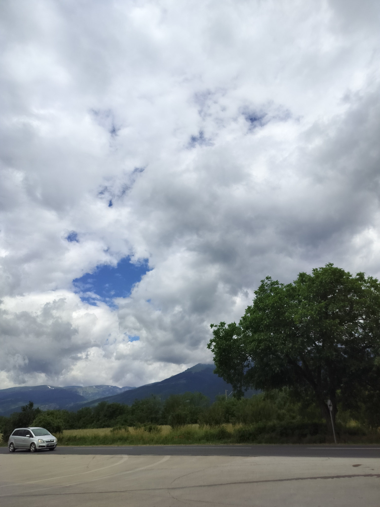 This is a landscape photograph taken in Tarnicheni, Bulgaria. The image captures a cloudy sky with patches of blue visible. In the foreground, there is a grey car parked on what appears to be a road or a parking area. To the right, a large tree with lush green foliage stands prominently. In the background, a mountain range is visible, with some areas appearing snow-capped. The overall environment suggests a natural, possibly rural, setting under an overcast sky.