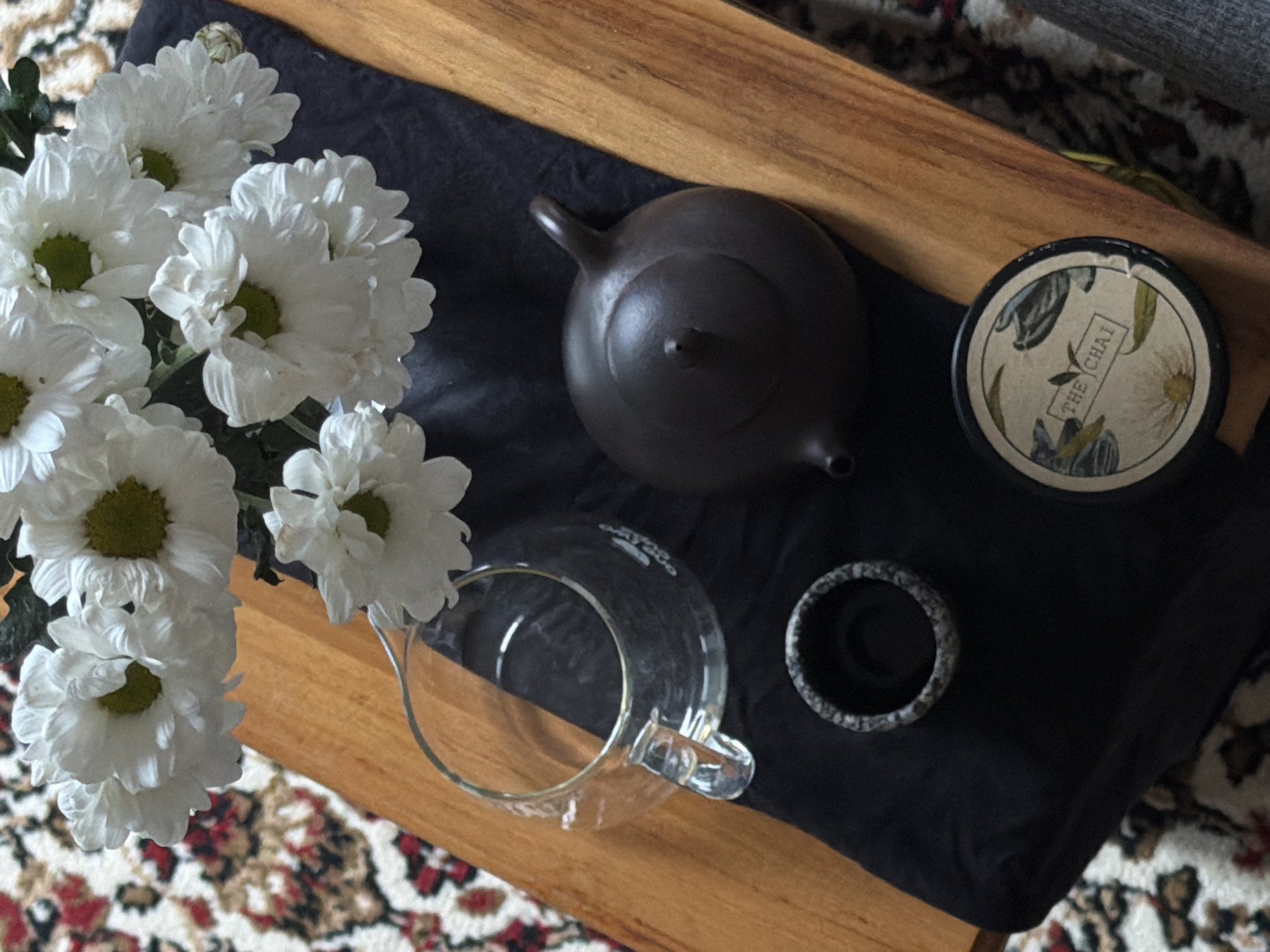 This overhead shot captures a serene tea setting arranged on a light brown wooden surface, partially covered by a dark, possibly black or navy, fabric mat. On the left, a cluster of white chrysanthemums with bright green centers adds a natural and fresh element to the scene.
To the right of the flowers, a dark brown, unglazed teapot, appearing to be made of clay or ceramic, sits on the fabric. Its traditional, minimalist design features a short spout and handle. Below the teapot, a clear glass pitcher or carafe, possibly made of borosilicate glass, is placed. Nearby, a small, dark, speckled gray-and-black cylindrical object, resembling a handleless tea cup or a tea pet holder, rests on the fabric. In the upper right corner, a round black container or coaster has a decorative lid featuring botanical illustrations of leaves and flowers in muted tones. The text "THE CHAI" is clearly visible on this lid within a rectangular frame.
The background reveals a section of a patterned rug with white, red, and dark brown motifs, suggesting an indoor setting. The lighting is soft and natural, indicating a peaceful daytime moment, possibly in North Kuta, Indonesia, as per the location context. There are no people visible, and the scene evokes a sense of calm and a ritualistic approach to enjoying tea.