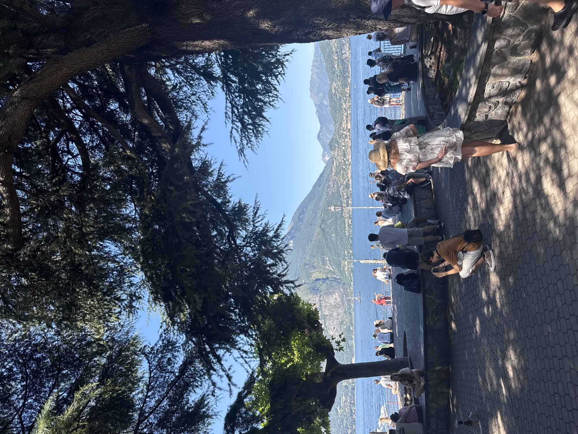 In Varenna, Italy, this sunny outdoor scene depicts a waterfront promenade lined with trees providing shade. In the foreground, a cobblestone path is visible, with dappled sunlight creating patterns on the ground. A young man in an orange t-shirt is squatting and taking a photograph of a duck that is walking on the path. Further back, a woman wearing a white floral dress and a straw hat stands with her back to the camera, facing the lake. Along the waterfront, a low stone wall separates the promenade from the lake, and a crowd of people are seated or standing, many facing the water. The lake itself is a deep blue, with distant mountains and buildings on the far shore. A sailboat is visible in the water. The overall impression is one of a popular tourist destination on a clear, warm day.