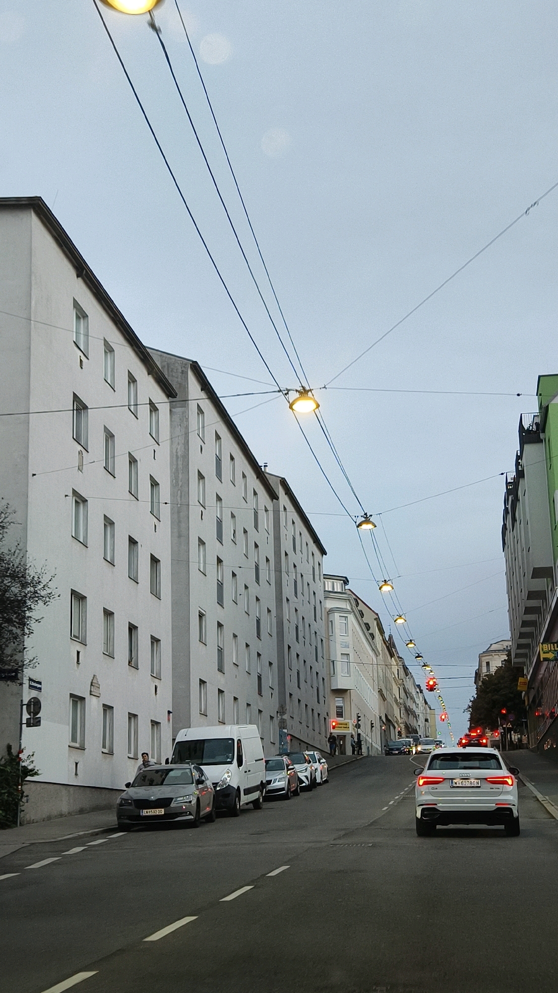 The image depicts a street scene in Vienna, Austria, characterized by a row of multi-story buildings on the left and a similar, though narrower, building on the right. The sky is overcast and grey. Overhead, a series of streetlights are suspended by wires, creating a pattern across the sky. The street itself is paved and shows lane markings.
On the left side of the street, parked cars are visible, including a white van and several other vehicles. A grey sedan with license plate "W-6592-DD" is in the foreground on the left. A person is standing near the driver's side door of this sedan.
The street slopes gently upwards and away from the viewer. Several cars are driving up the street, including a white Audi SUV in the right lane, displaying license plate "W-6278-QN". Traffic lights are visible further up the street, some of which are illuminated red. People are seen walking on the sidewalks in the distance. The buildings are uniformly designed with multiple windows. Some commercial signage is visible on the right side, including a yellow sign with "BILL" in black letters.