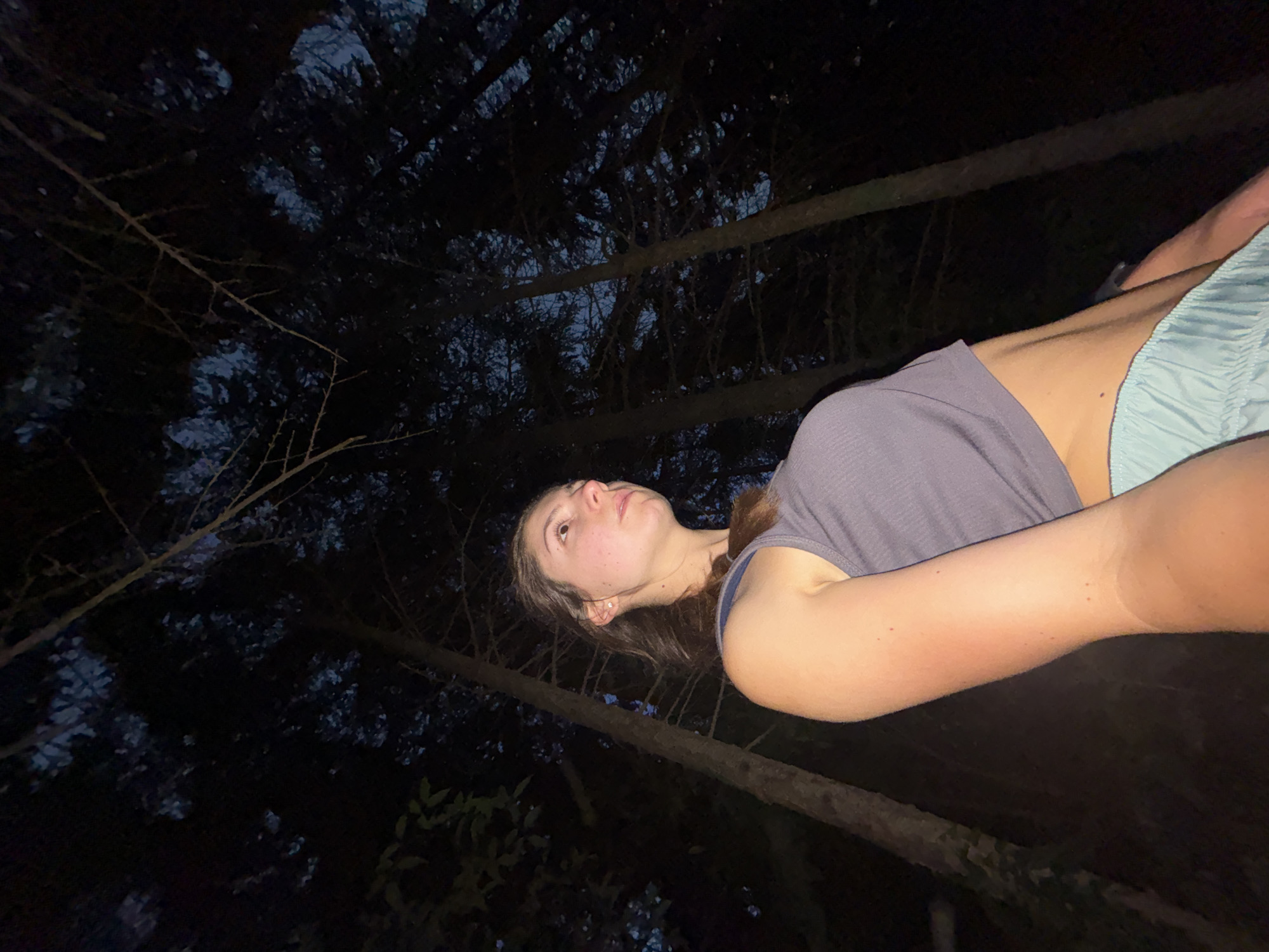The image depicts a low-angle shot of a young woman standing in a forest at night. She is wearing a grey tank top and light blue shorts. Her hair is brown and pulled back. The background is dominated by tall trees, their branches silhouetted against a dark sky. The lighting is dim, suggesting natural light from the moon or artificial light sources outside the frame. There are no identifiable landmarks or structures that would place this image specifically in Varna, Bulgaria.