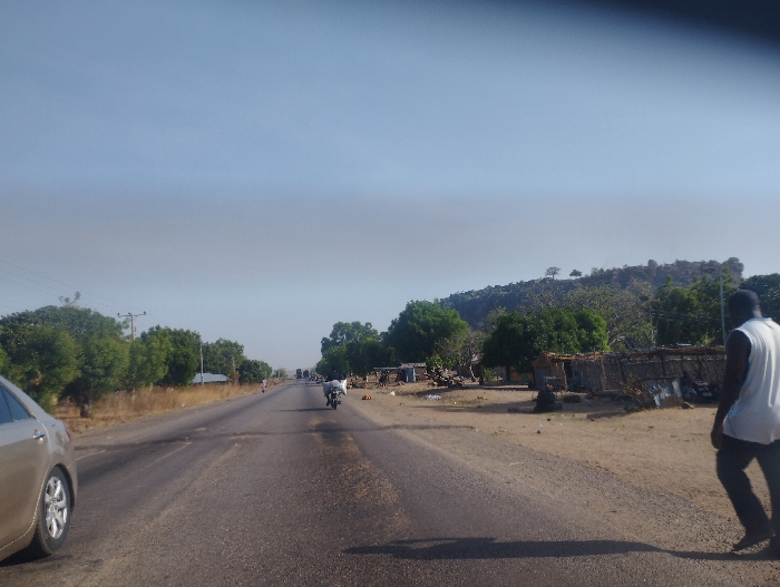 A paved road, seemingly a highway, cuts through a sparsely populated area. The road is empty except for a vehicle in the foreground and two motorbikes further down the road, one of which has a rider. The road is framed by a grassy shoulder on both sides. There are trees on the edge of the road, and a rocky hill in the distance, possibly a mountain. The sky is blue and clear. A single man walks toward the left side of the image, away from the road, at a slight angle. He is wearing a white sleeveless shirt and dark pants. Behind him, a collection of lean-to structures are constructed of metal, wood and thatch. There are more trees behind the structures, some of which are in the background on the left, and most of which are clustered at the foot of the hill. The photograph appears to have been taken from a vehicle traveling on the road, looking straight ahead. The photo was taken in Ture-Balam, Nigeria, at the coordinates 9.797 latitude and 11.382 longitude.