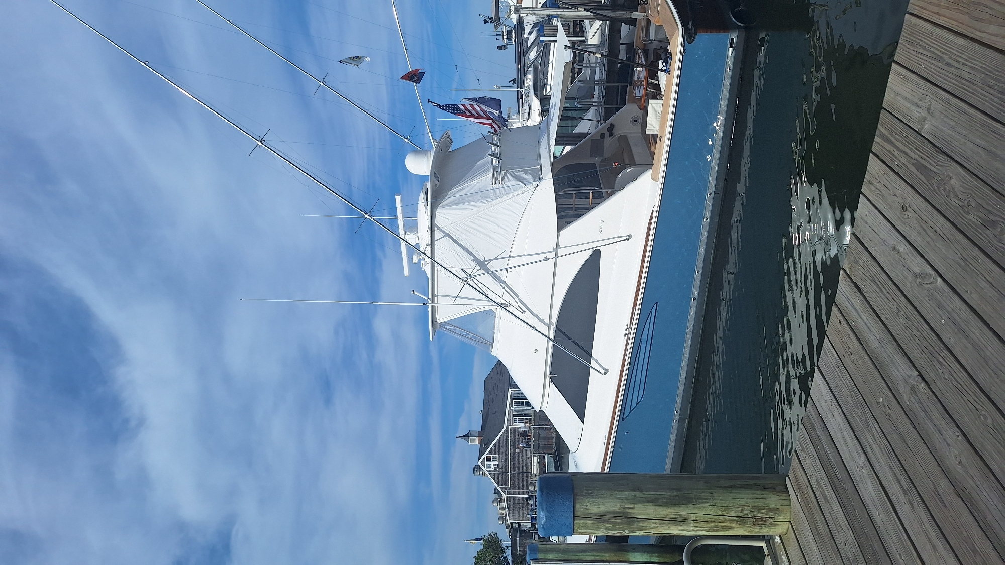 A large, white and blue sportfishing yacht is docked at a pier on a sunny day with a cloudy sky. The yacht has a prominent white superstructure with a clear enclosure. The hull is painted a deep blue with a thin white stripe. Fishing outriggers extend from the top of the superstructure. An American flag and another blue flag are flying from the yacht. In the background, several other boats are moored and a cluster of buildings with a distinctive steeple, characteristic of Nantucket's architecture, are visible. The foreground shows a weathered wooden pier and the dark, reflective water of the harbor. The scene appears to be in Nantucket, Massachusetts, United States of America, judging by the architectural style of the buildings. There are no people clearly visible in the immediate foreground or on the yacht.