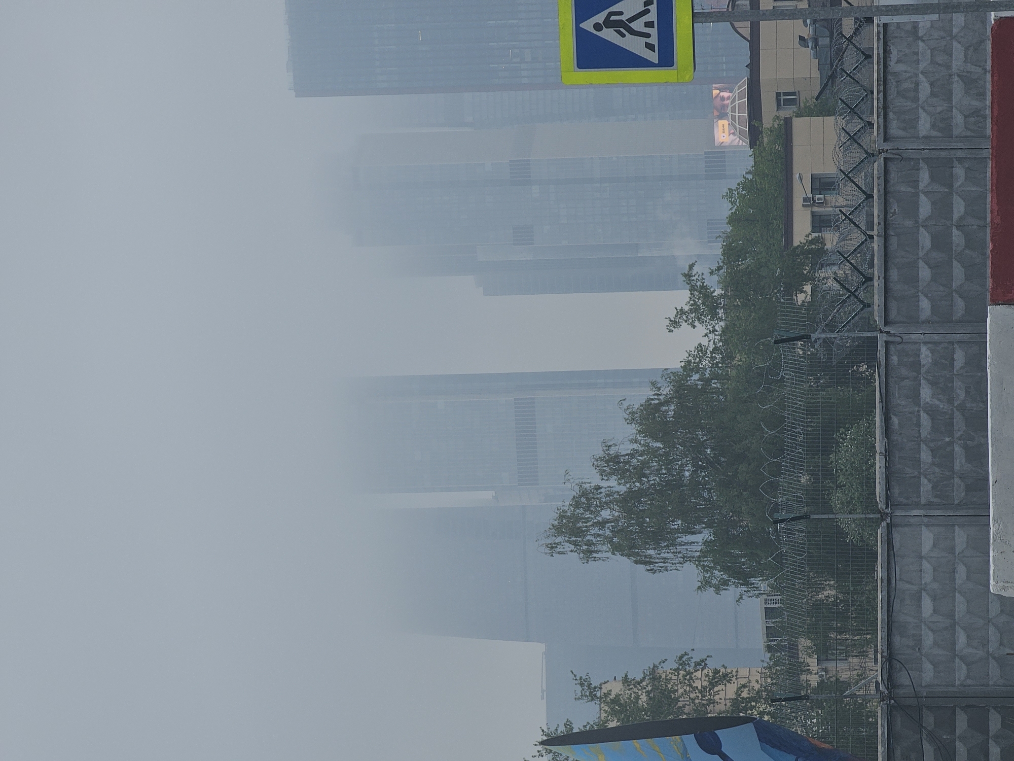 The image shows a view of the Moscow International Business Center (Moscow-City) through dense smog or fog. The skyscrapers are partially obscured by the haze. A pedestrian crossing sign is visible in the upper right corner. A concrete wall topped with barbed wire runs along the right side of the frame, and trees are present between the wall and the distant buildings.