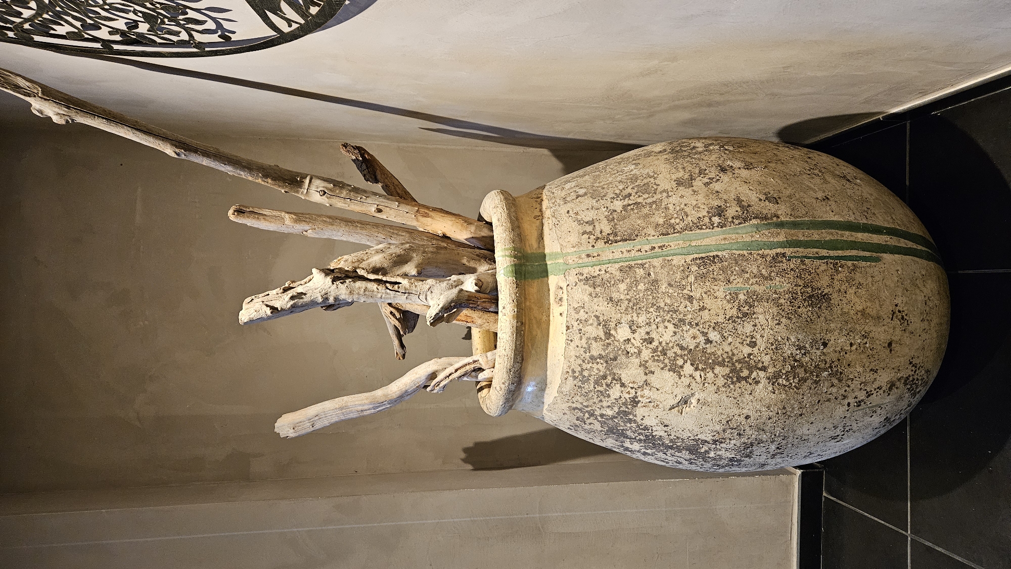 A large, weathered terracotta pot filled with dry branches and twigs stands in a corner against a textured, neutral-toned wall. The pot has a rounded base and a wide opening, with green streaks running down its exterior. The branches are of varying sizes and textures, creating a rustic display. The floor is tiled in black. In the background, a partial view of a decorative, black, circular wall hanging with a leafy motif is visible. The lighting creates shadows on the wall, suggesting an indoor setting. The location is an interior space in L'Île-Rousse, France.
