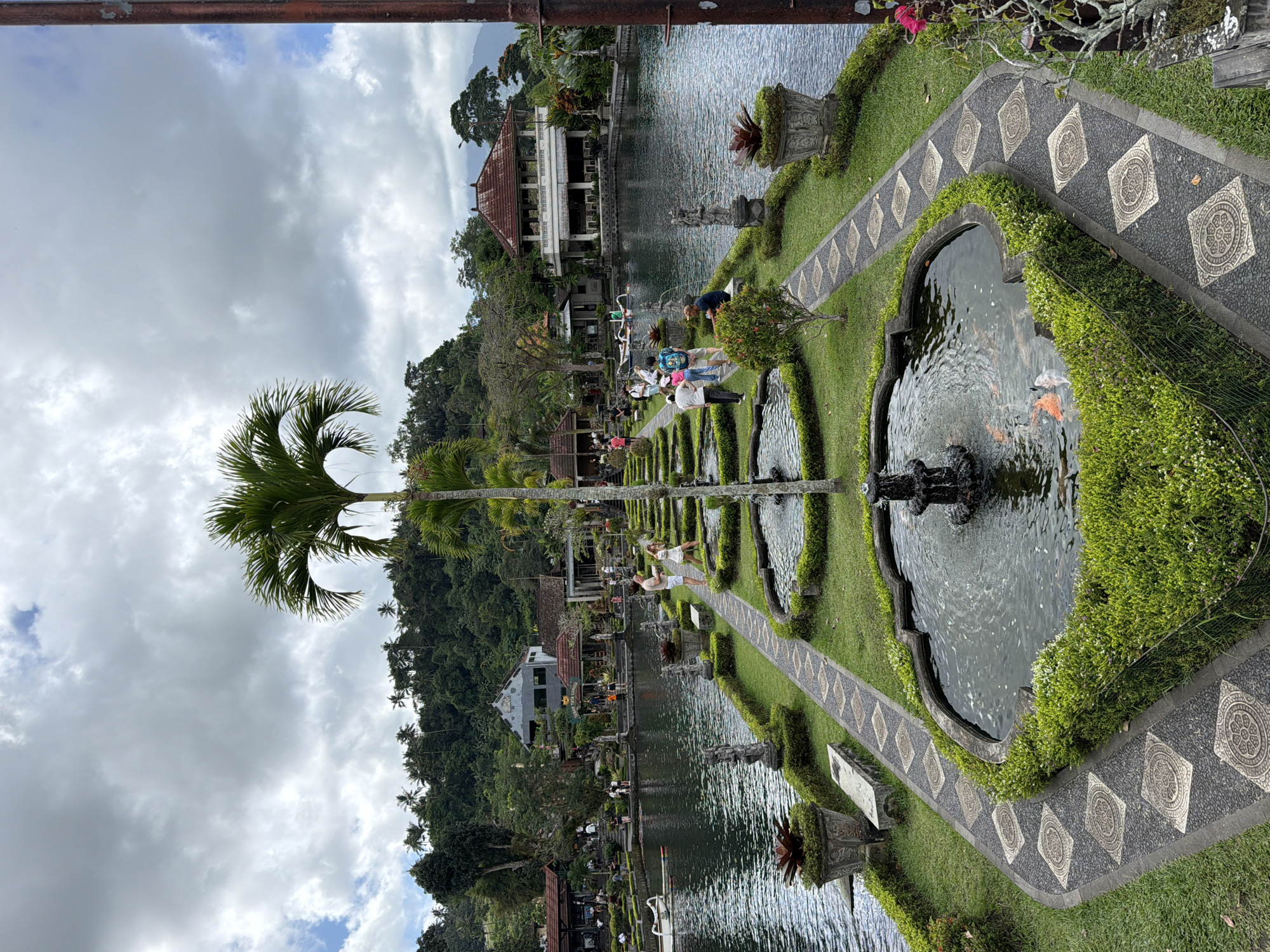 This is a vertical, eye-level shot of the Tirta Gangga water palace in Abang, Indonesia. The foreground features a formal garden with a central pathway of patterned stones leading to a series of ornamental ponds with fountains. Lush green hedges border the pathways and ponds. In the foreground pond, several koi fish are visible swimming.
A prominent palm tree stands near the center of the pathway, rising towards the sky. In the mid-ground, people are strolling along the pathways and near the large body of water. Several buildings with traditional Indonesian architecture, including a prominent two-story structure with a red tiled roof, are visible on the far side of the water. The background is dominated by a lush, green hillside covered in trees and vegetation, with mountains faintly visible in the distance. The sky is overcast with dramatic, grey clouds. The overall scene conveys a sense of tranquil beauty and historical significance.