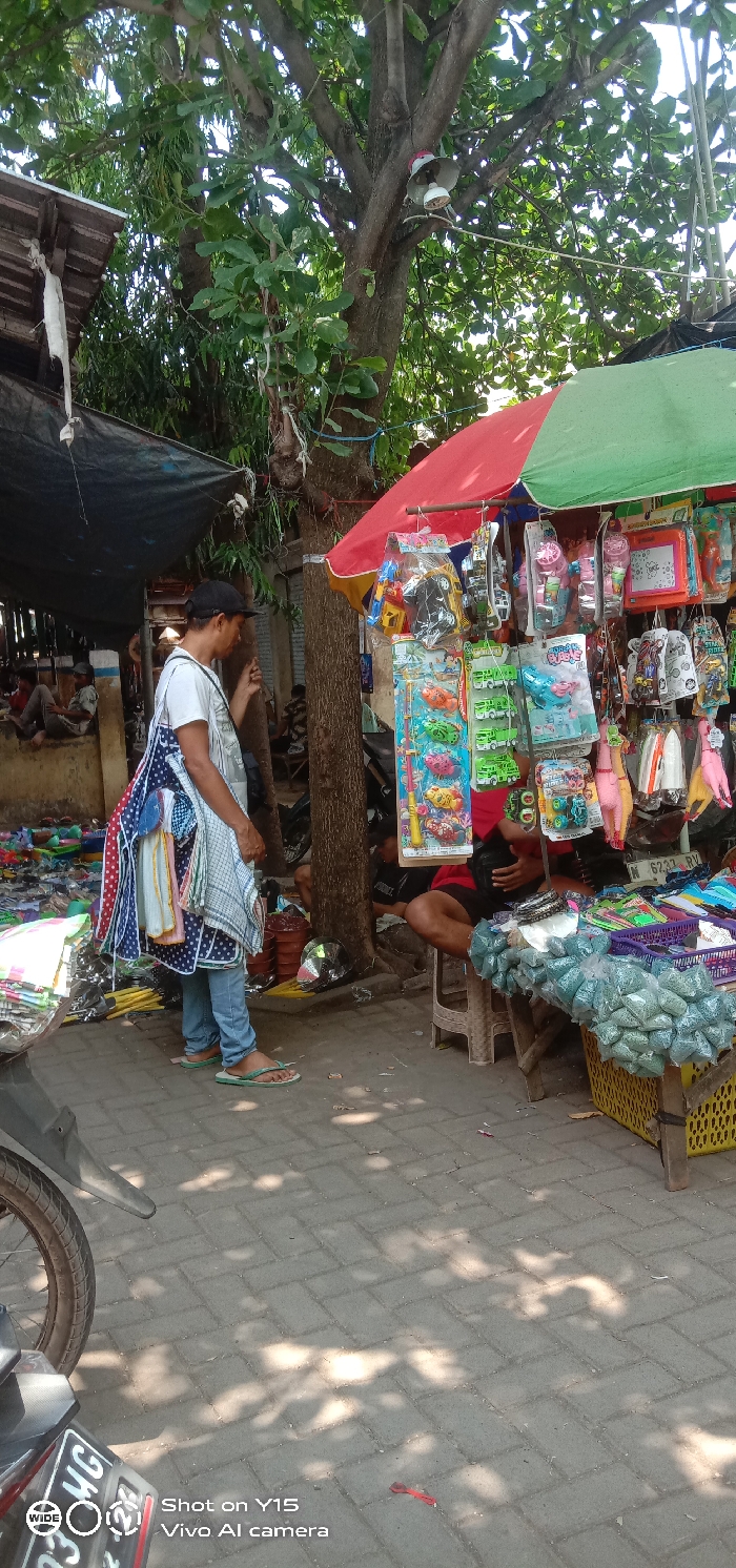 This image displays an outdoor market scene in the city of Maron, Indonesia, under bright daylight. In the left-center, a male subject is standing, wearing a white t-shirt, blue jeans, a black baseball cap, and light green flip-flops. He carries multiple patterned fabric items, including aprons and towels, draped over his left shoulder and arm. His right hand is positioned near a tree trunk.
To the right of the standing subject, a market stall is active beneath a large, two-toned umbrella (red and green sections). The stall features numerous packaged items, predominantly toys such as vehicles, fishing kits, bubble blowers, rubber chickens, and drawing boards, hanging from its framework. Partially visible behind this stall, a second male subject, wearing a dark shirt and shorts, is seated on a light-colored plastic stool, attending to the merchandise. The stall's display includes a yellow plastic crate containing multiple clear plastic bags filled with small, light green objects, and a purple plastic basket holding various goods.
A large tree with dense green foliage stands centrally, its trunk serving as an anchor for the market stall's umbrella via a visible blue rope. Two horn-style speakers are affixed to a branch of this tree in the upper right quadrant of the image. In the blurred background on the left, several other individuals are visible near makeshift structures covered with dark tarpaulins. The ground is paved with gray interlocking concrete pavers, displaying a pattern of sunlit areas and shadows cast by overhead trees and structures. A portion of a dark-colored motorcycle, including its rear wheel and a partial license plate with the characters "MG," is visible in the lower left foreground.