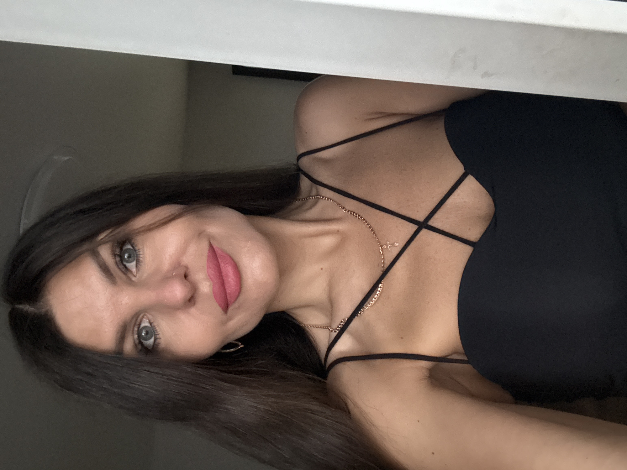A woman with dark brown hair and blue eyes is looking at the camera, with pink lipstick on. She is wearing a black top with thin straps and a rose gold necklace with a small cross pendant. Her left shoulder is partially obscured by a white vertical object, possibly a door frame. The background is dark and out of focus, suggesting an indoor setting. The image appears to be a selfie. The location of Chaikovsky, Russia is not depicted in the image.