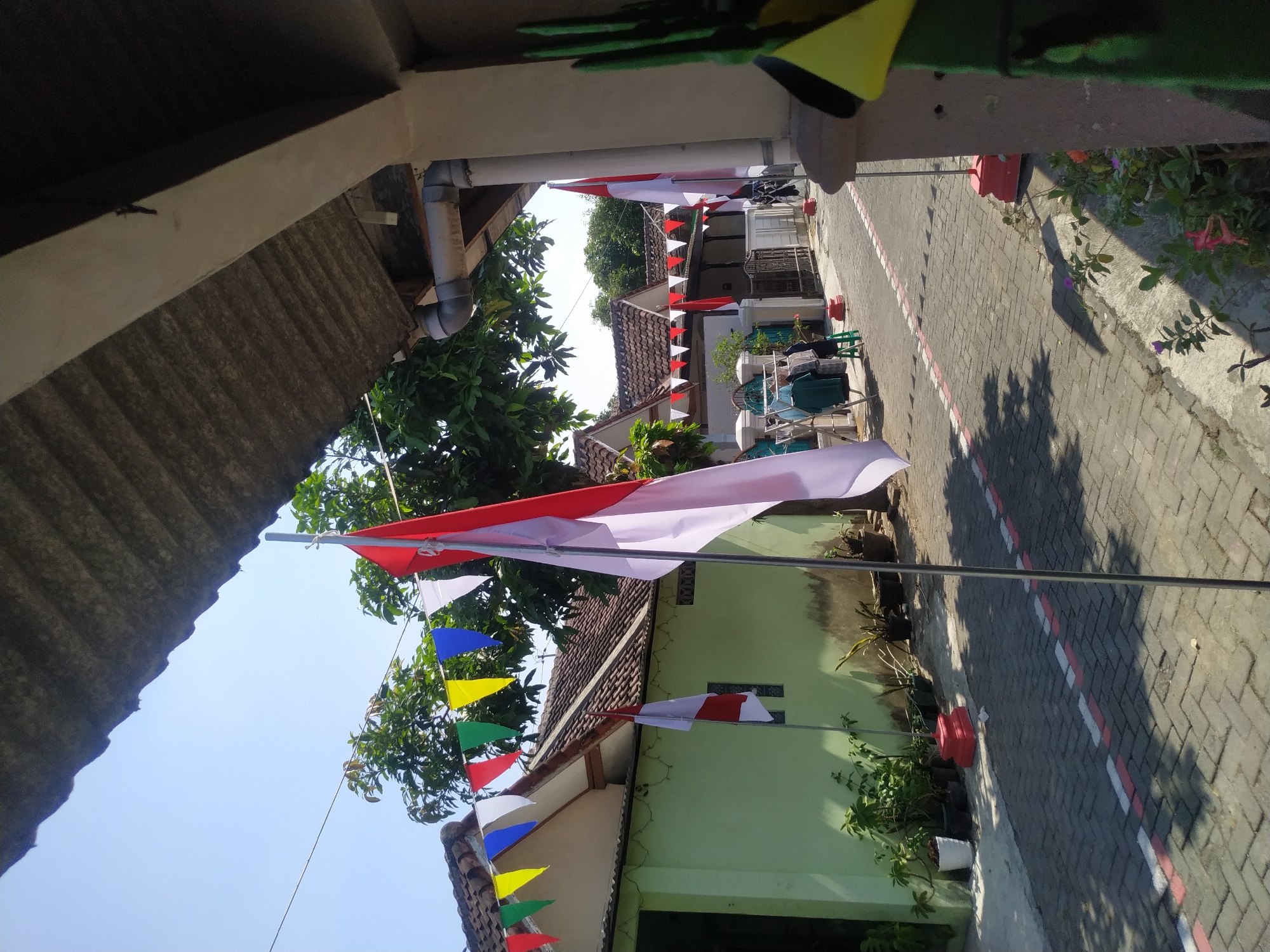 In Banyudono, Indonesia, a narrow paved street is adorned with Indonesian flags and colorful triangular pennant banners, suggesting a celebration or national holiday. The street is lined with single-story houses with terracotta roofs and light-colored walls, some painted in pale green. Trees with lush green foliage are visible between the houses. On the left side of the street, potted plants are placed near a green-painted house. A clothesline with several garments hangs in front of a house further down the street. Two Indonesian flags, one larger and more prominent in the foreground, are mounted on poles with red bases. The ground is paved with interlocking bricks, marked with intermittent red and white painted lines. The sky is clear and blue, indicating a sunny day. The scene appears to be residential, with no individuals actively engaged in the frame.