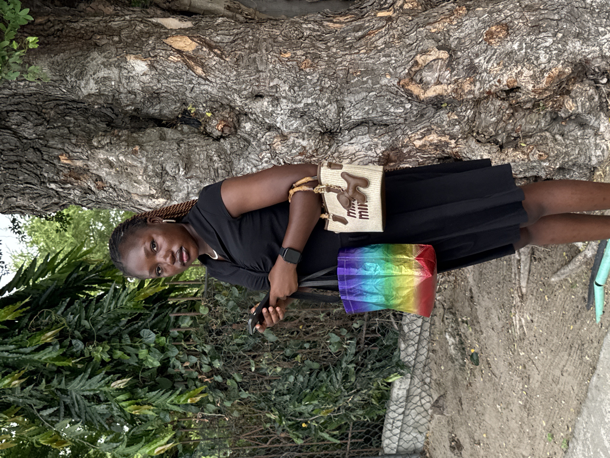 The image features a young Black woman standing outdoors, looking directly at the viewer with a neutral to slightly smiling expression. She is wearing a black dress or top and skirt, with a dark-colored watch visible on her left wrist.
The woman is carrying two bags. Over her left shoulder, she has a light-colored, possibly straw or woven, shoulder bag featuring brown leather accents. The brand name "miu miu" is clearly visible in brown text on this bag. In her right hand, she holds a dark object, likely a phone, and also carries a vibrant rainbow-colored tote bag, which appears to be made of a crinkled or textured fabric.
The background consists of a large, textured tree trunk to her left, and dense green foliage, including bushes and vines, behind and to her right. A metal fence is partially visible amidst the greenery, separating the plants from the ground, which appears to be a mix of dirt and concrete.
The setting suggests an outdoor urban or semi-urban environment, consistent with the location of Lekki, Nigeria. The natural lighting indicates it is daytime, with soft, diffused light suggesting either an overcast sky or an area with significant shade, as there are no harsh shadows. The overall scene captures a moment of the woman posing casually in a natural yet defined outdoor space.