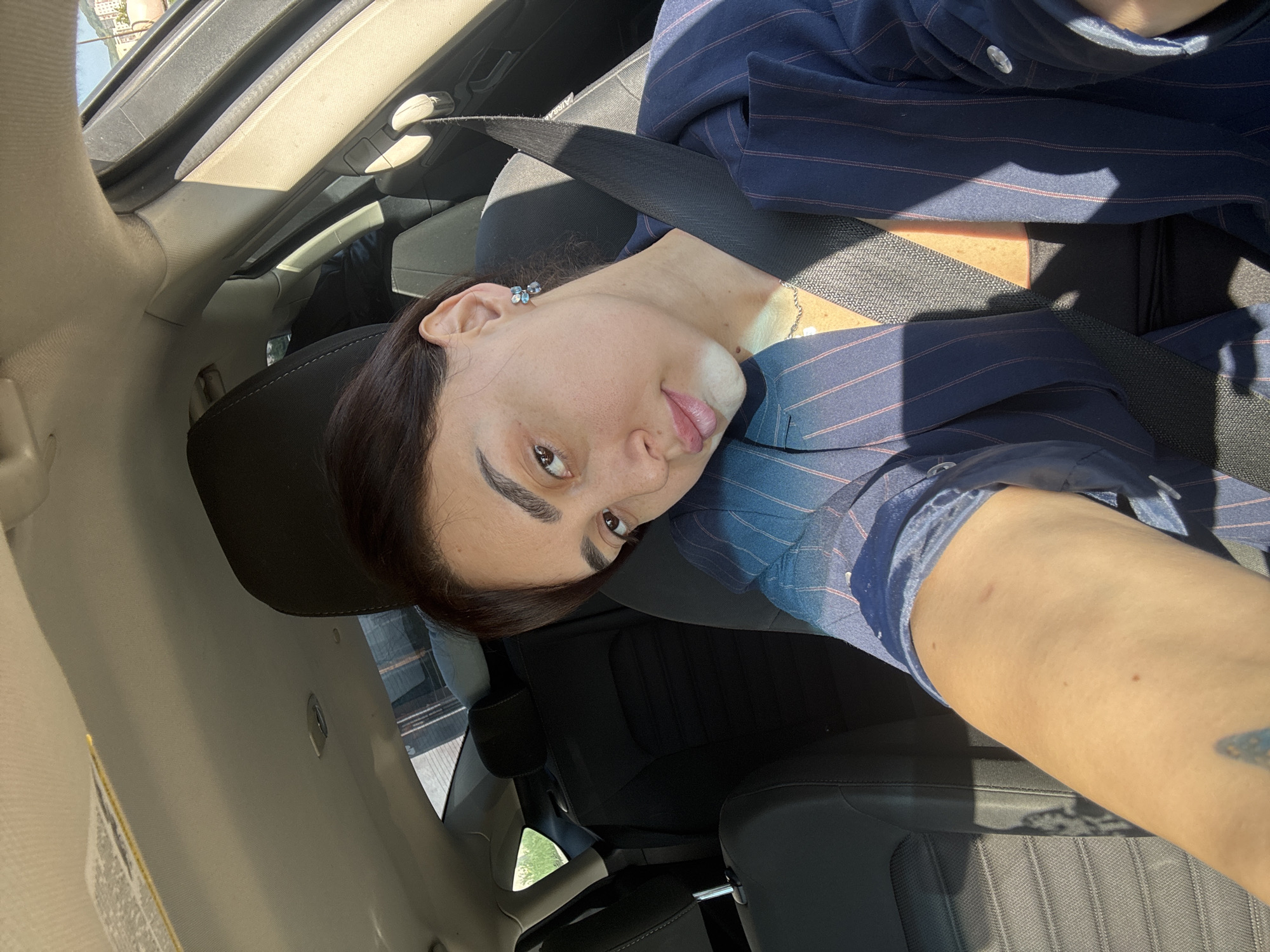 A selfie taken inside a car. A woman with brown hair is in the driver's seat, wearing a blue pinstripe blazer and a seatbelt. The interior of the car is visible. The image was taken on a sunny day. The location is likely Tbilisi, Georgia, based on street views visible through the car window.