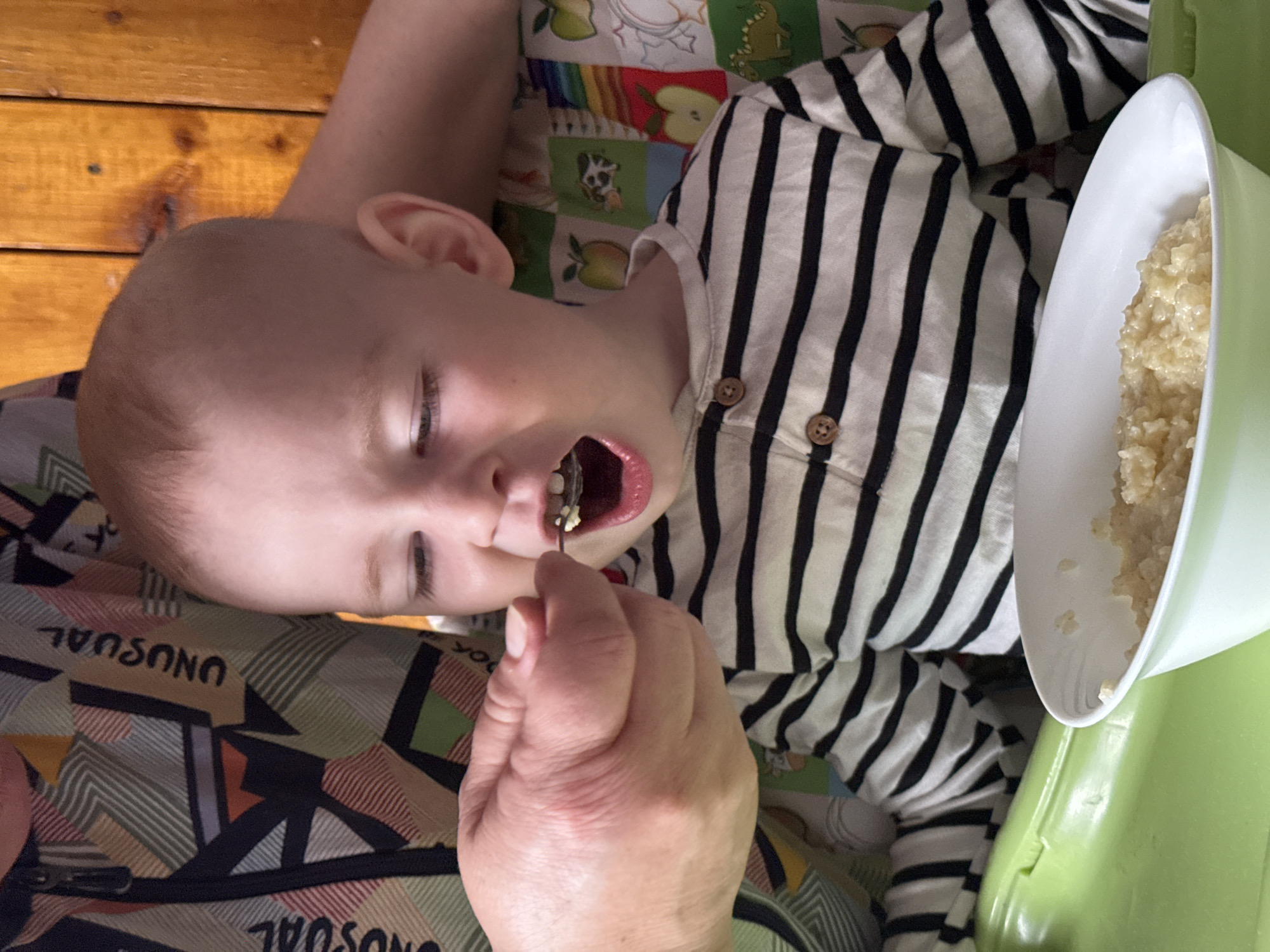 A baby lies on someone's lap, wearing a striped shirt. An adult hand is seen feeding the baby with a spoon from a white and green bowl filled with cooked rice. The background includes a wooden surface, a colorful patterned fabric, and a green highchair. The location cannot be determined from the image and is not Уральское, Russia.
