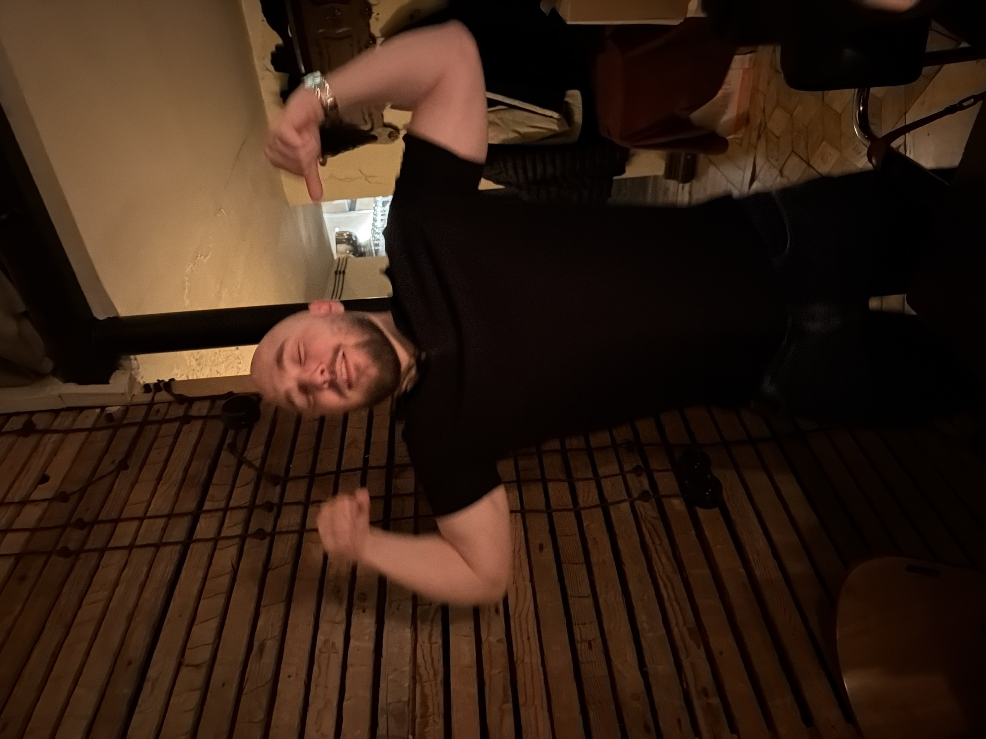 The image depicts a bald, bearded adult male, wearing a black polo shirt and dark trousers, standing indoors under dim, warm lighting. He is centrally framed, posing with both arms bent, flexing his biceps, eyes closed, and a slight smile. A metallic wristwatch is present on his left wrist. The immediate background on the left consists of a wall constructed from horizontal wooden planks, overlaid with dark, thin vertical elements and a dark, circular fixture. A light-colored wooden chair back is partially visible in the lower left. To the right, a doorway leads into an adjacent space where a metallic vessel and multiple stacked glass objects are discernible. The walls in this area are light-colored and textured, with a corresponding light-colored ceiling. Dark fabric is draped over objects in the lower right, and the floor exhibits patterned tiling. The precise location within the city of Tbilisi, Georgia, cannot be determined from the visual information provided.