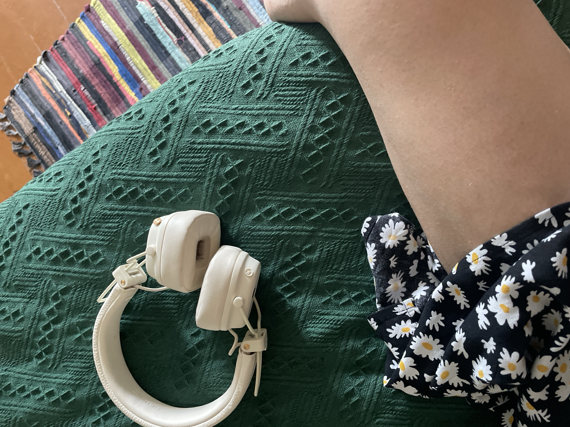 A close-up, overhead shot shows a pair of cream-colored headphones resting on a textured, dark green blanket. To the right of the headphones, a person's bare leg is visible, with goosebumps dotting the skin. A black fabric with a white and yellow daisy pattern is draped across the lower left corner of the image, partially obscuring the leg. A colorful, striped rug is positioned in the upper right corner, partially visible behind the blanket. The scene appears to be indoors, on what looks like a couch or bed, with no specific city or location identifiable.