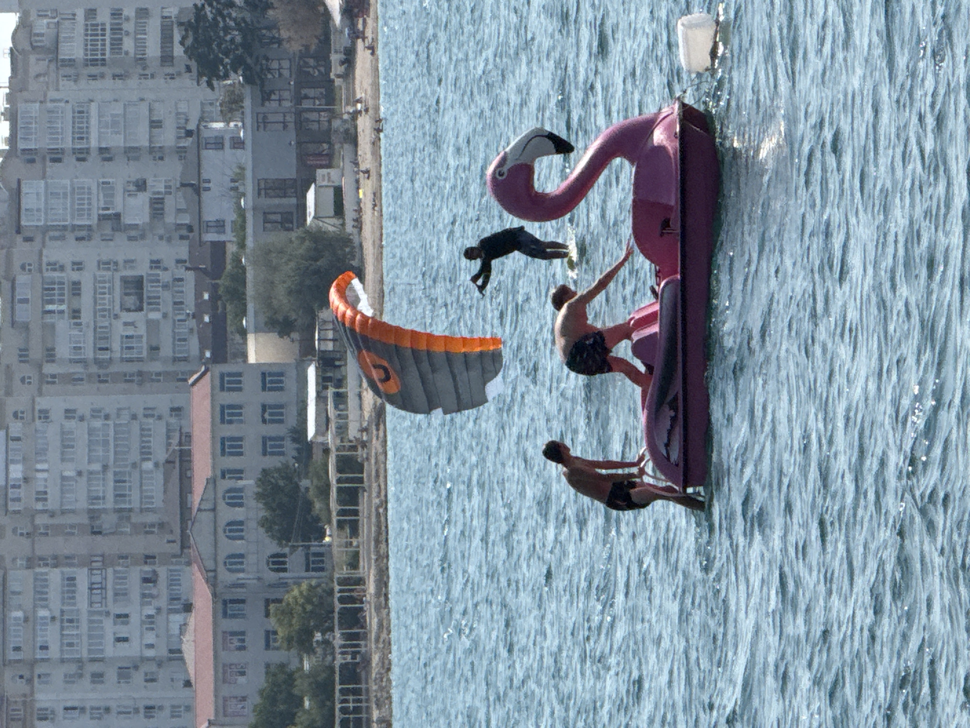 In Sevastopol, Ukraine, a bright pink flamingo-shaped paddleboat is partially submerged in the choppy blue water of the sea. Three individuals are on or near the paddleboat. One person, wearing black shorts, is kneeling on the paddleboat, reaching out towards the water. Another person, wearing black swim trunks, is standing on the edge of the paddleboat, holding onto a railing. A third person is in the water behind the paddleboat, appearing to be kiteboarding, with a large, orange and grey kite in the air above them. The paddleboat is tethered to a white buoy on the right side of the frame. In the background, there is a sandy beach with people and umbrellas, followed by a row of white, arched structures and further back, a densely populated area of multi-story buildings. The overall scene suggests a recreational activity taking place on a sunny day.
