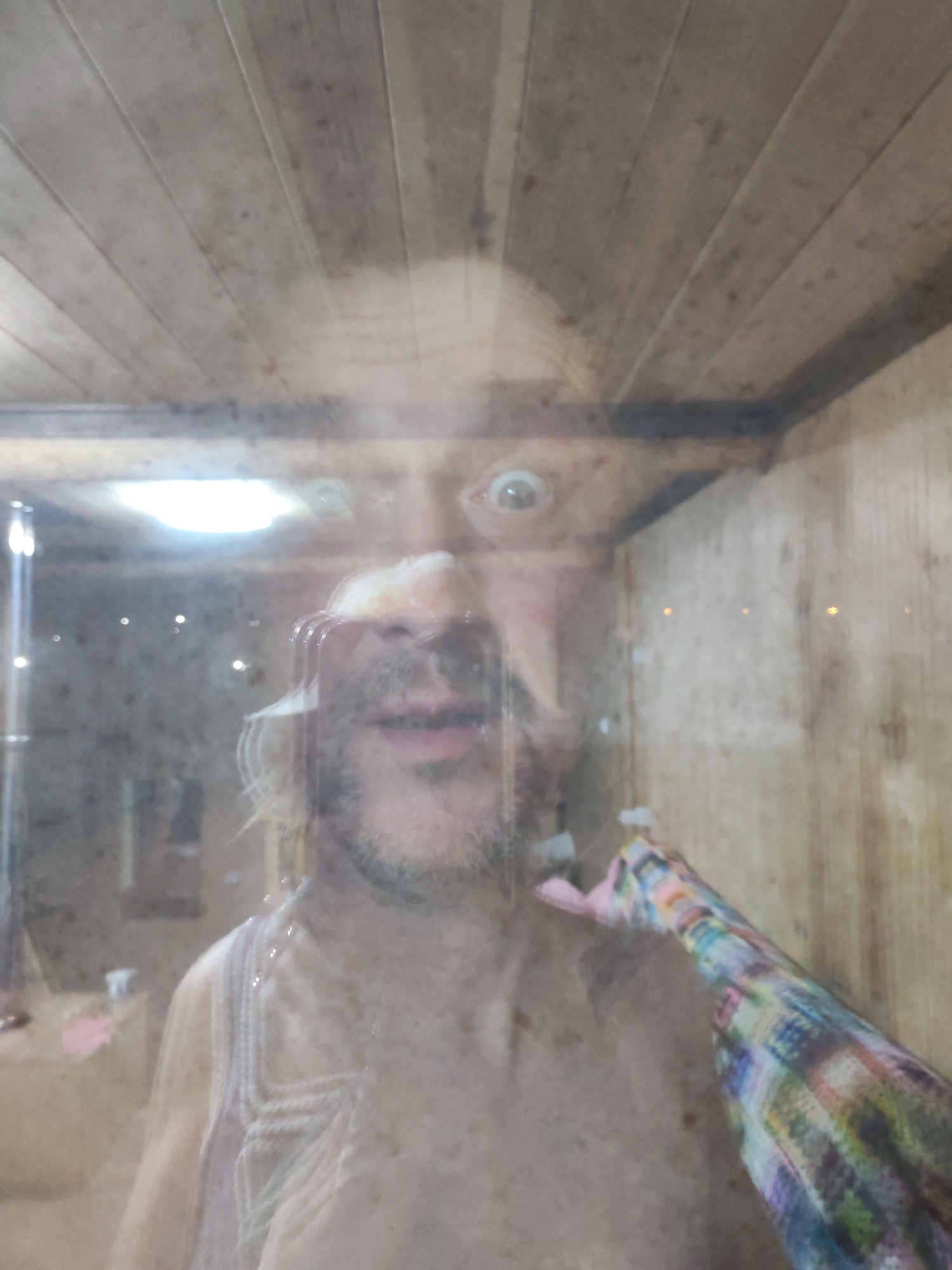 The image appears to be a selfie taken in a sauna, possibly located in Chaikovsky, Russia, based on the provided information. A man with light skin, a beard, and wide eyes is visible, seemingly distorted by condensation or reflections on the sauna's glass or plastic enclosure. The sauna has wood paneling on the ceiling and walls. A colorful, patterned towel is visible on his right side, possibly being held by him or another person. Some objects are visible in the background, but difficult to identify due to the distortion and reflections.