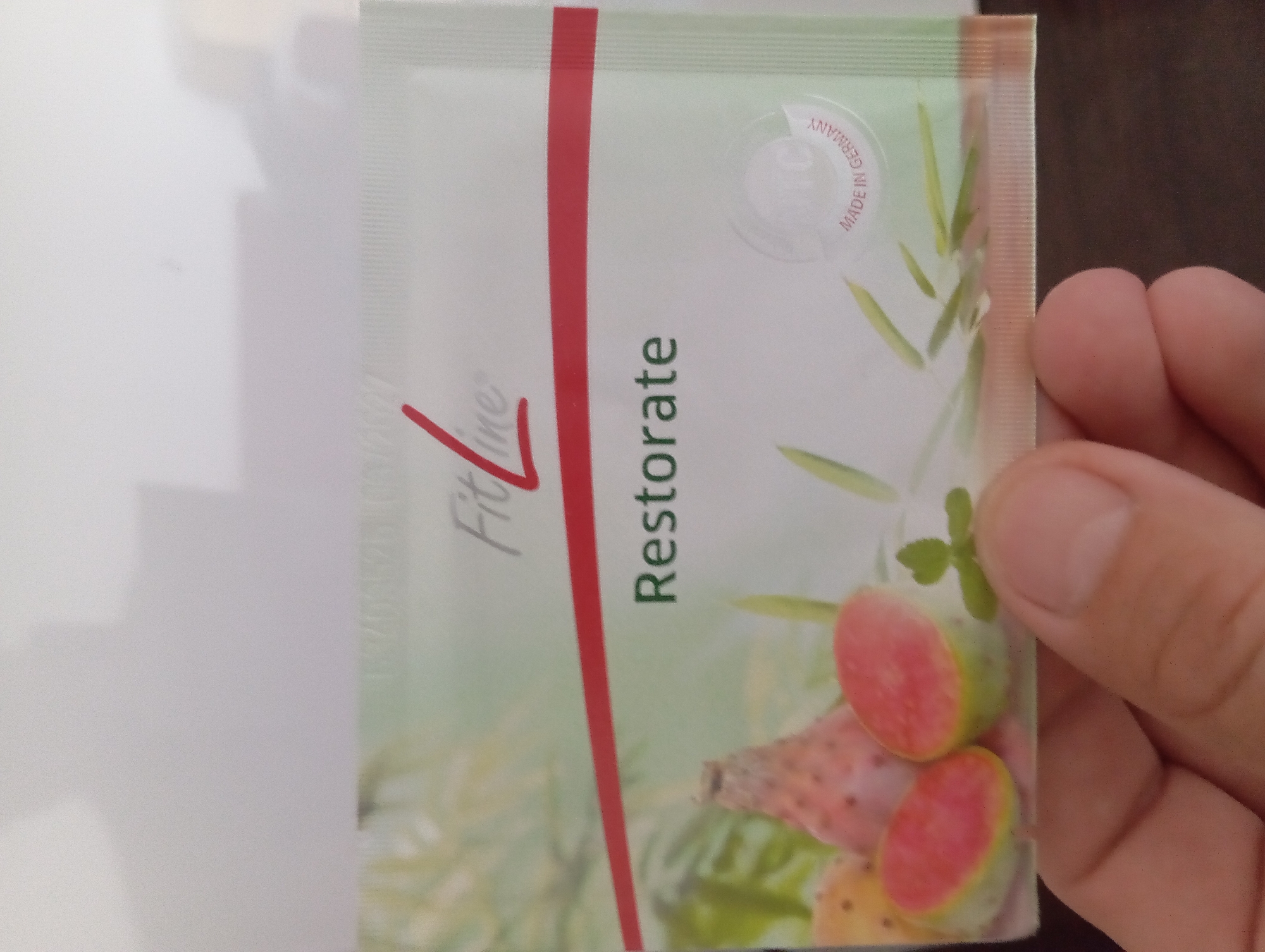 A hand holds a small, rectangular packet with a white background. The packet features a red stripe across the top half, beneath which is the word "Restorate" in green text. To the left of "Restorate" are images of prickly pears, some whole and one halved to reveal its pink interior. To the right of "Restorate" is a circular emblem with the text "Pm-International" and "Made in Germany" around the perimeter. The packet is positioned over a dark surface, and the background is a light, blurred color. The image was taken in Heitersheim, Germany.