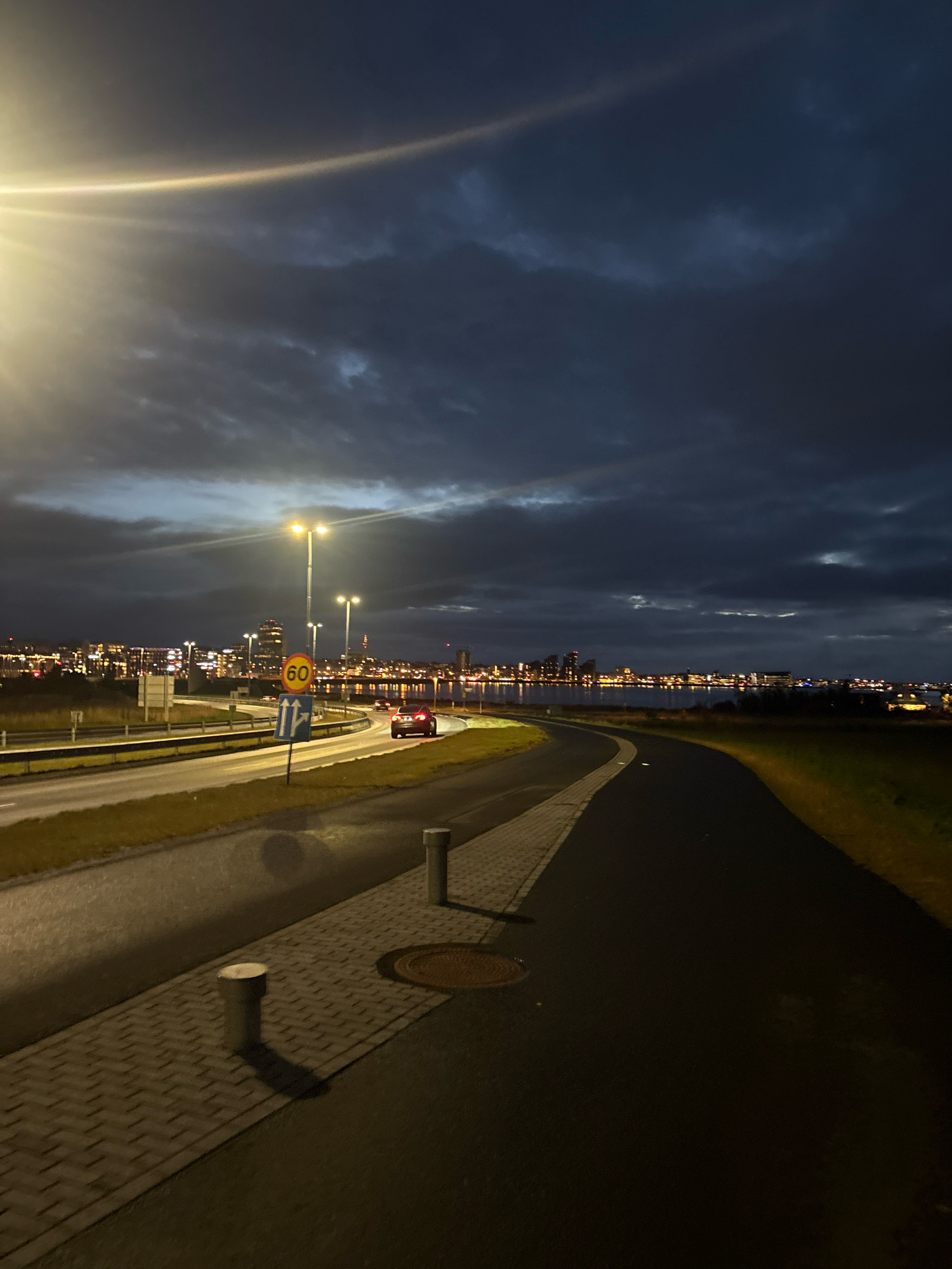 The image depicts a street scene at dusk in Reykjavik, Iceland. A road curves to the right, with a pedestrian pathway bordering it. Streetlights illuminate the road and pathway, casting long shadows. Traffic signs, including a speed limit sign of 60, are visible. A dark-colored car is driving on the road. In the distance, the Reykjavik skyline is visible, with numerous buildings lit up, indicating an urban setting. The sky is dark and cloudy, with a bright light source, possibly a street lamp or car headlight, creating a lens flare in the upper left corner. The environment appears to be coastal, with water visible in the middle ground between the road and the city. The overall impression is of an evening in a city with active infrastructure and a dramatic sky.