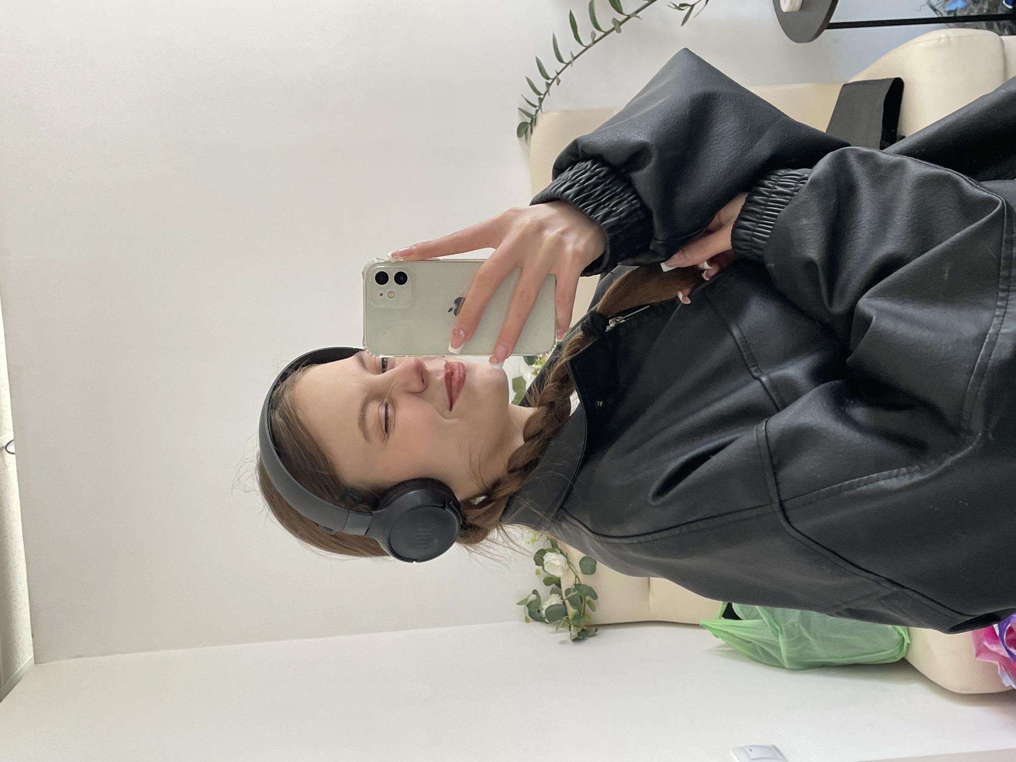 A woman is taking a selfie with a smartphone. She is wearing black headphones and a black leather jacket, and has her hair in a braid. The background appears to be a room with white walls. The location of Фоки, Russia cannot be determined from the image.