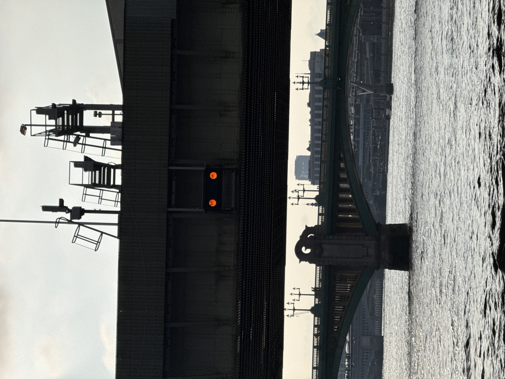 This image shows a view under the Tower Bridge in London, United Kingdom, with the River Thames sparkling in the sunlight. A portion of the bridge’s steel structure is visible on the left, featuring a signal with two illuminated orange lights. On the right, part of the bridge's architectural details including the tower, arch, and lampposts is seen. Beyond the bridge structure, the skyline of London with several buildings is visible.