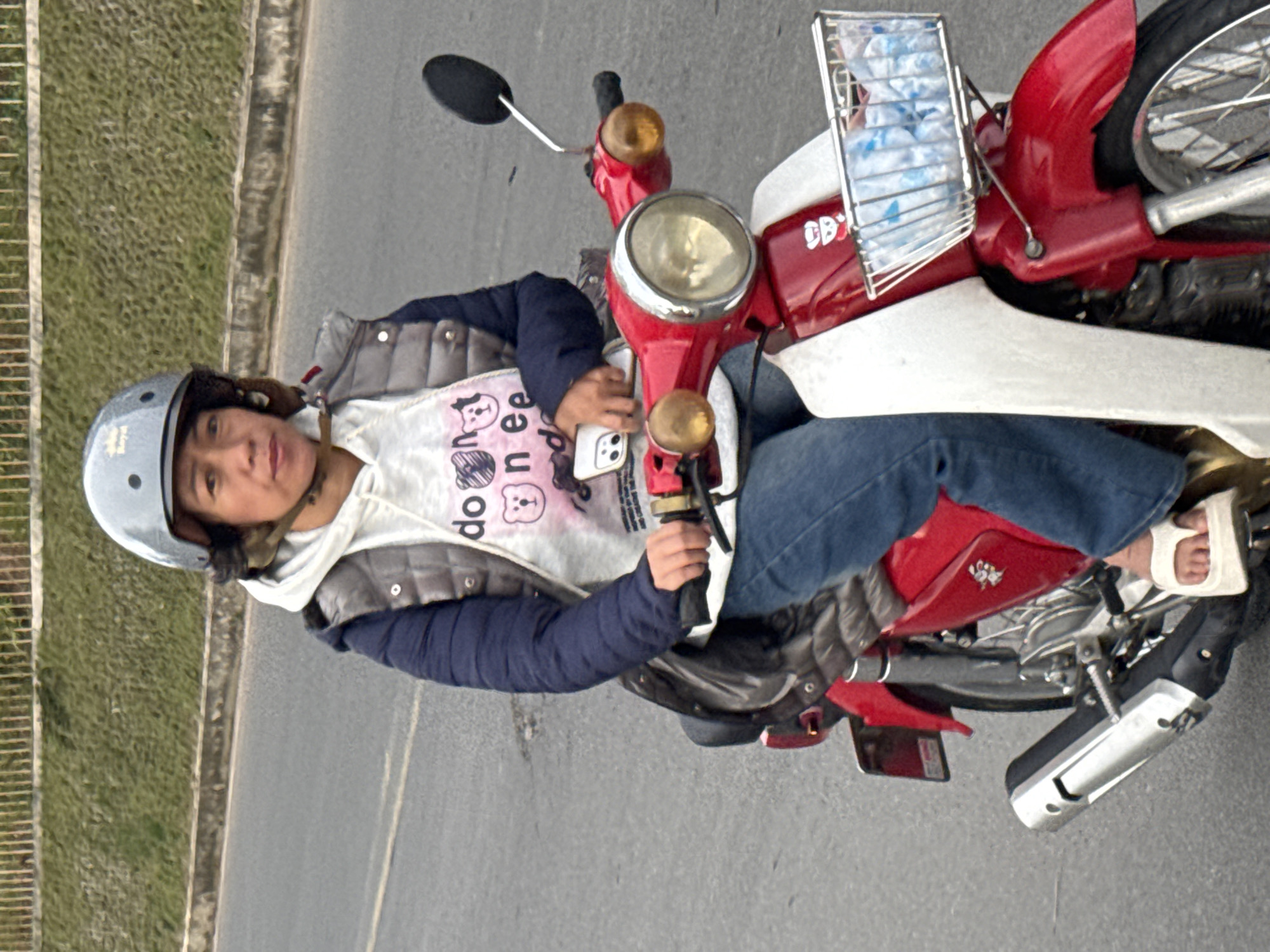 A woman wearing a gray helmet, a gray puffer vest over a white hoodie with pink graphics, and blue jeans rides a red and white motorcycle on a paved road. The motorcycle has a silver basket on the front containing plastic water bottles. The woman is holding a white smartphone in her right hand. The background shows a grassy embankment and a fence. The scene appears to be in Da Lat, Vietnam, a city known for its French colonial architecture and temperate climate.