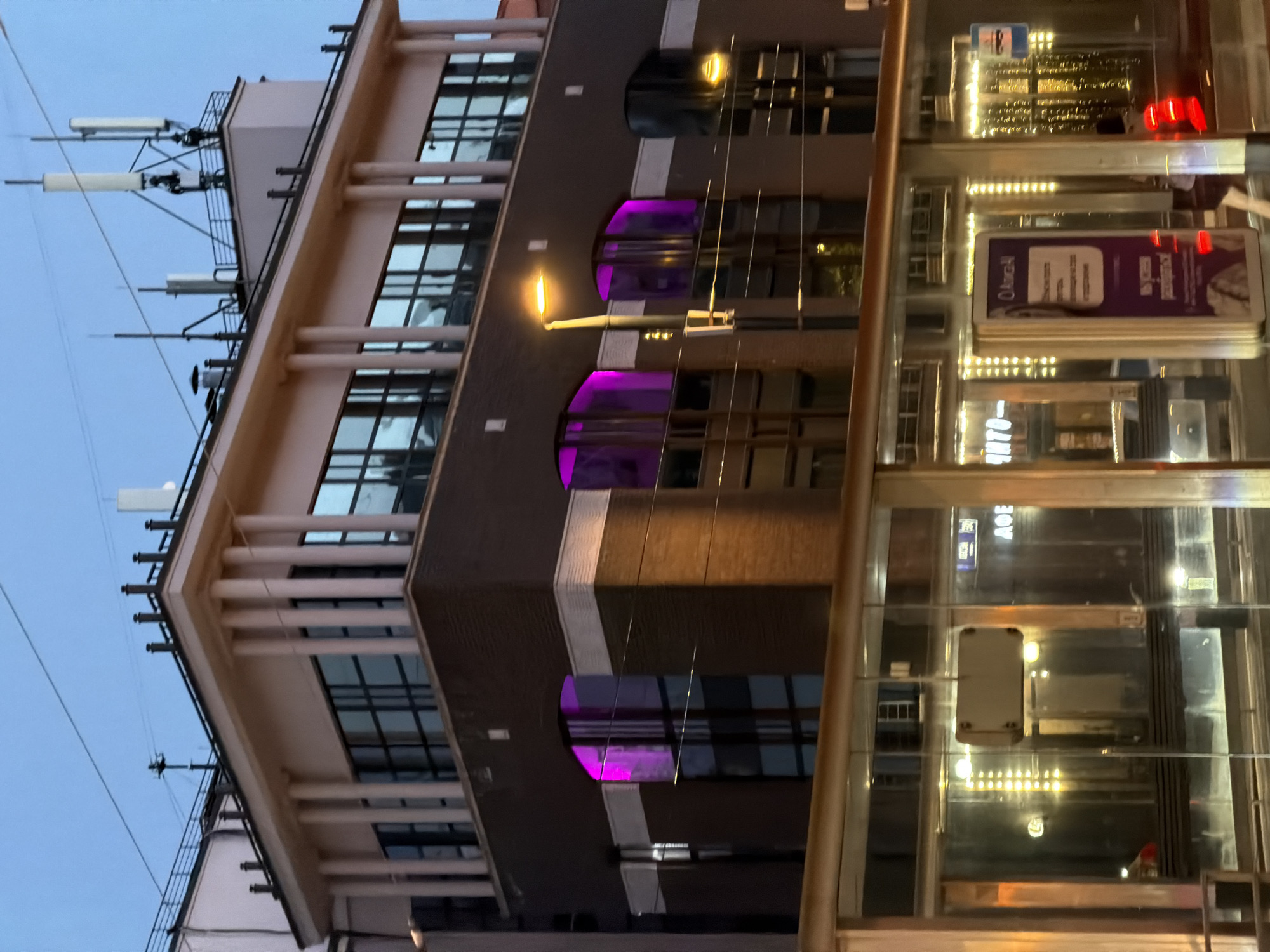 Here's a description of the image:
The image shows the "Izvestia Hall" building in Moscow, Russia. This building has a distinctive architectural style, featuring a combination of modern and classic elements. The facade of the building has a series of arched windows illuminated with purple light. The upper part of the building has a series of antennas and satellite dishes. The building is adjacent to a glass-encased structure that seems to be an elevator. A street lamp is visible in the foreground. The overall impression is that the photo was taken at dusk or twilight.