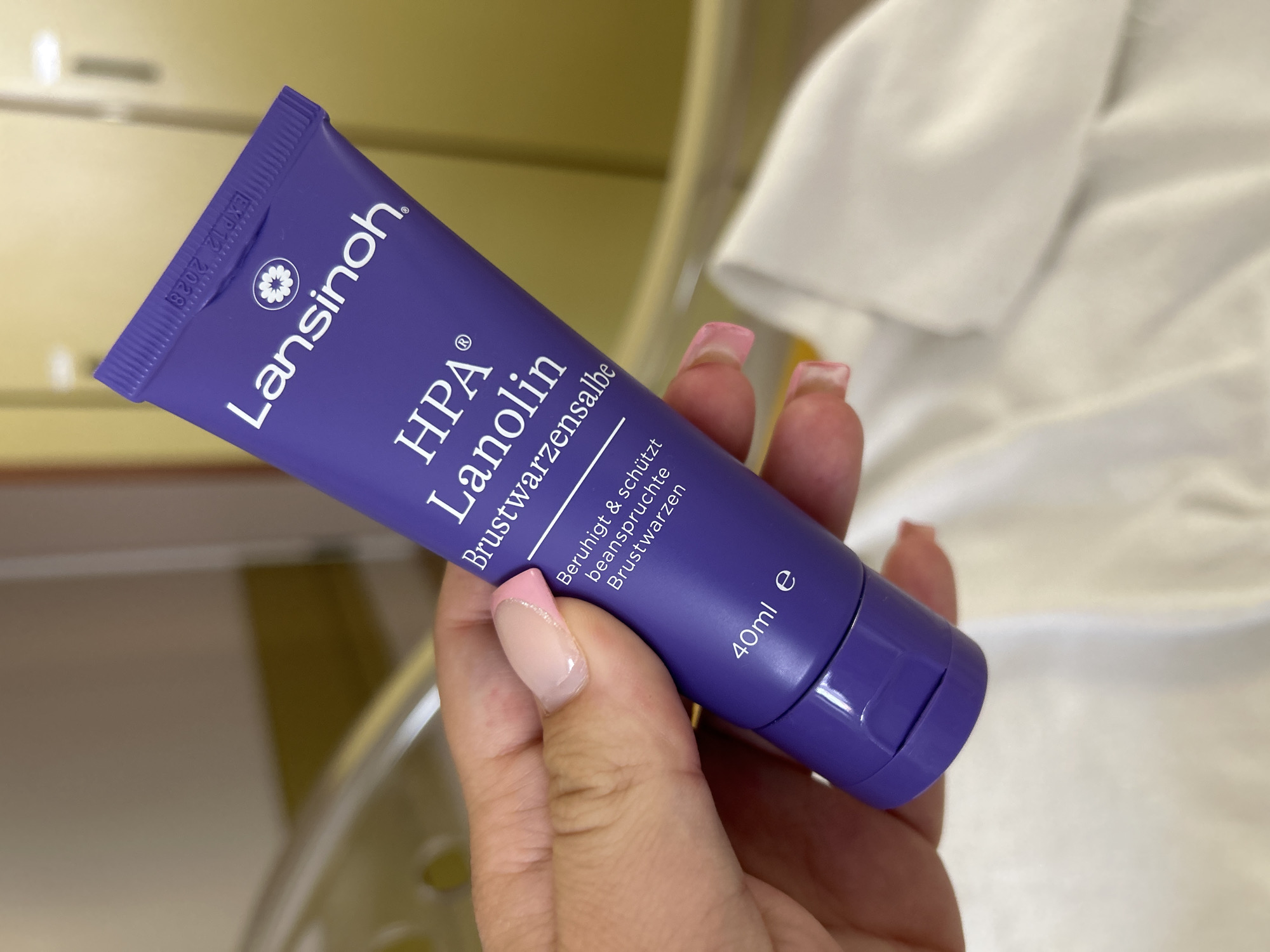 A hand with French manicured nails holds a purple tube of Lansinoh HPA Lanolin Brustwarzen (nipple balm) which has a white logo and text. The tube is angled and illuminated. The background is blurred, showing what appears to be a pale yellow wall and the edge of a white sheet or fabric. There is no discernible information about the location in Gummersbach, Germany.