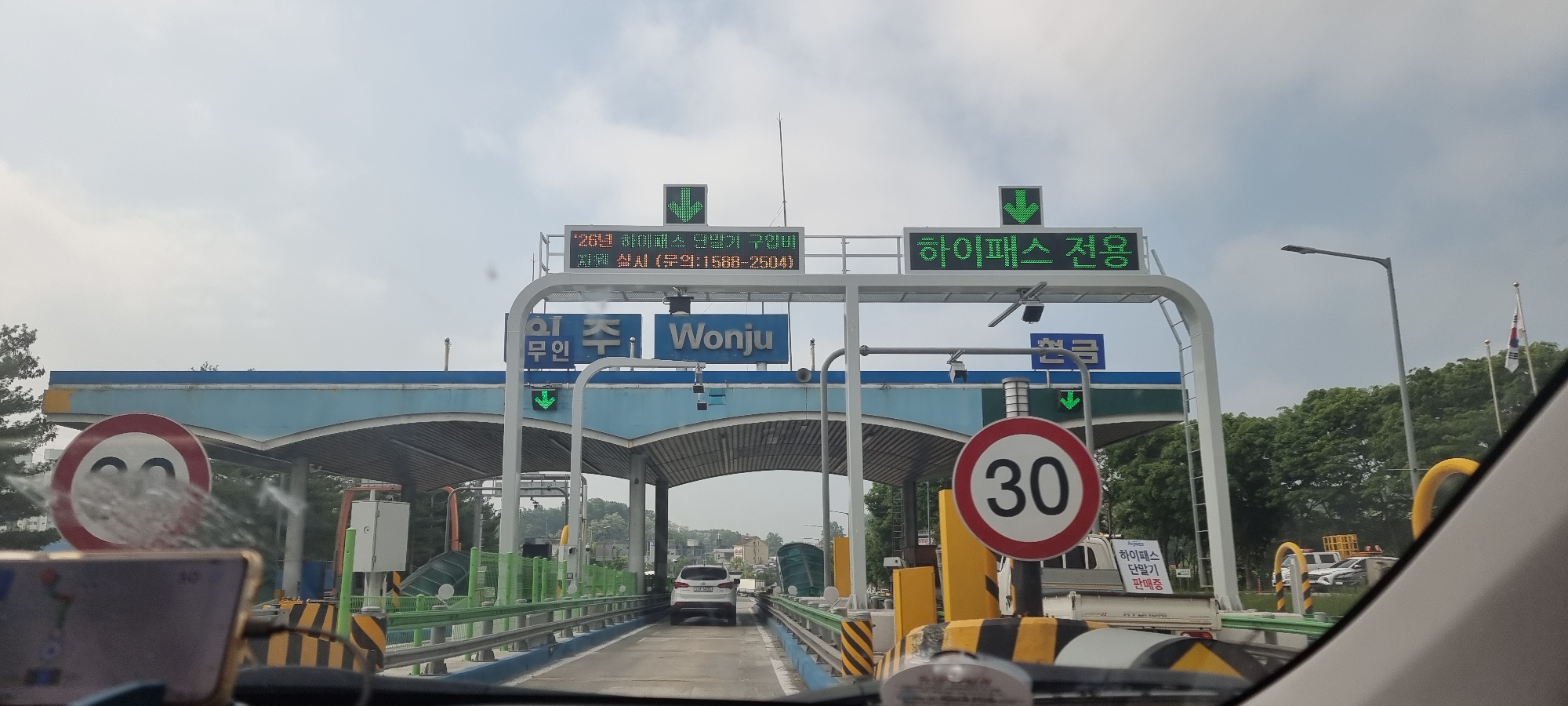 The image shows a vehicle approaching a toll plaza on a road in Wonju, South Korea. The toll plaza has lanes designated for "Hi-Pass," an electronic toll collection system. A green LED sign displays Korean text and a green downward arrow over each lane. The sign over the regular toll lane also includes the text "'26년 하이패스 단말기 구입 지원 실시(문의: 1588-2504)". Signs indicate that unmanned tolls are available. A speed limit sign with "30" is visible on the right. There is also a sign stating "하이패스 단말기 판매중", which translates to "Hi-Pass Terminal Selling". A South Korean flag is visible in the background on the right. Other vehicles are present in the plaza. The photo was taken from inside a vehicle, with part of the dashboard and a navigation device visible in the foreground.