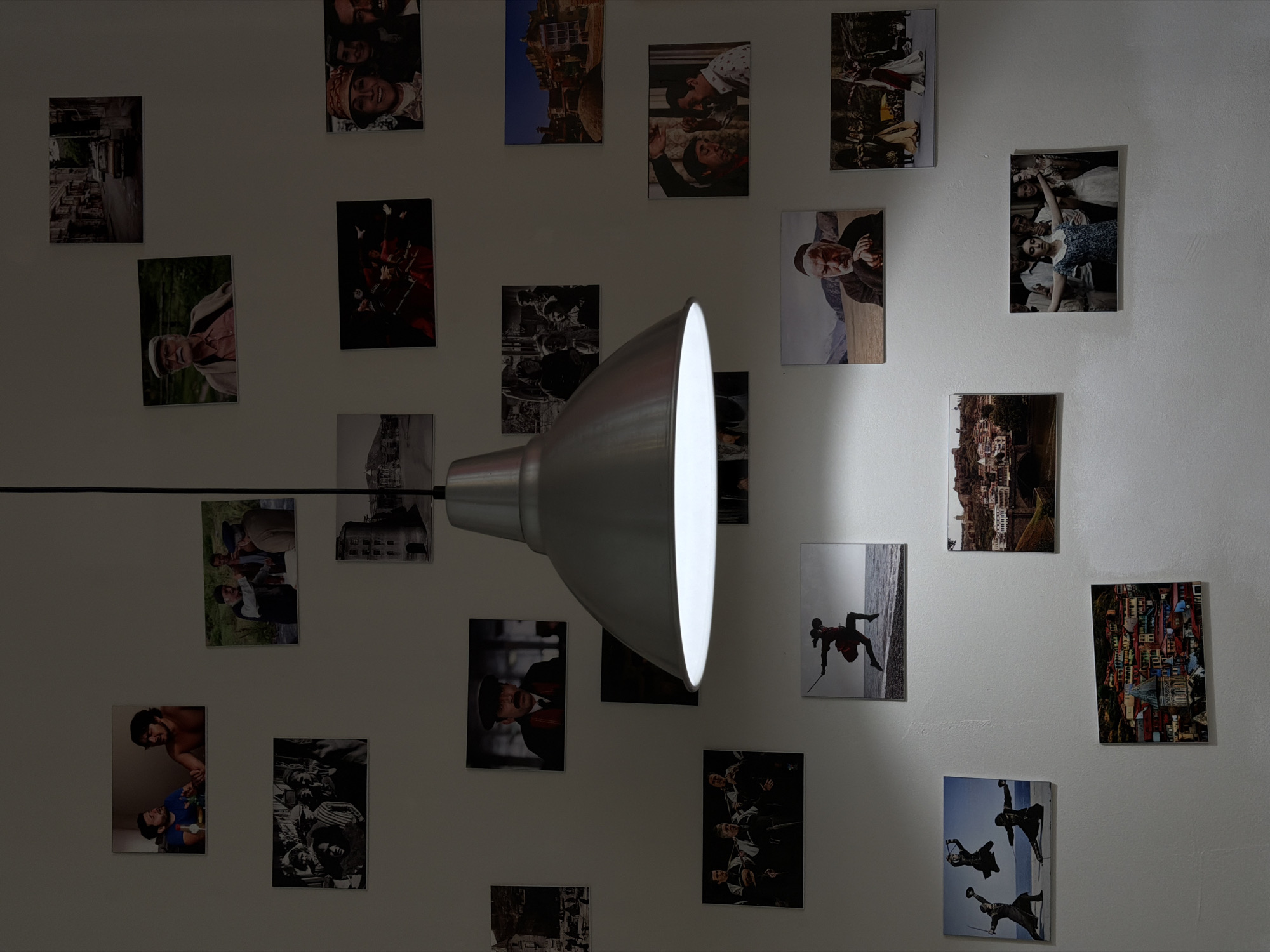 A medium shot depicts a white wall adorned with an assortment of photographs, with a modern lamp providing illumination. The photos are mounted in a seemingly random arrangement, displaying diverse subjects ranging from portraits to landscapes and scenes of people. The lamp, positioned in the center of the frame, casts a spotlight on a section of the wall, creating a contrast between the illuminated area and the rest of the photos. The wall itself is plain and serves as a neutral backdrop for the displayed photos and the lamp. The scene evokes a sense of a personal space, possibly a home or a studio, where the photographs serve as a form of decoration and expression. The location is not discernible from the image, and there is no indication of it being in Moscow, Russia.