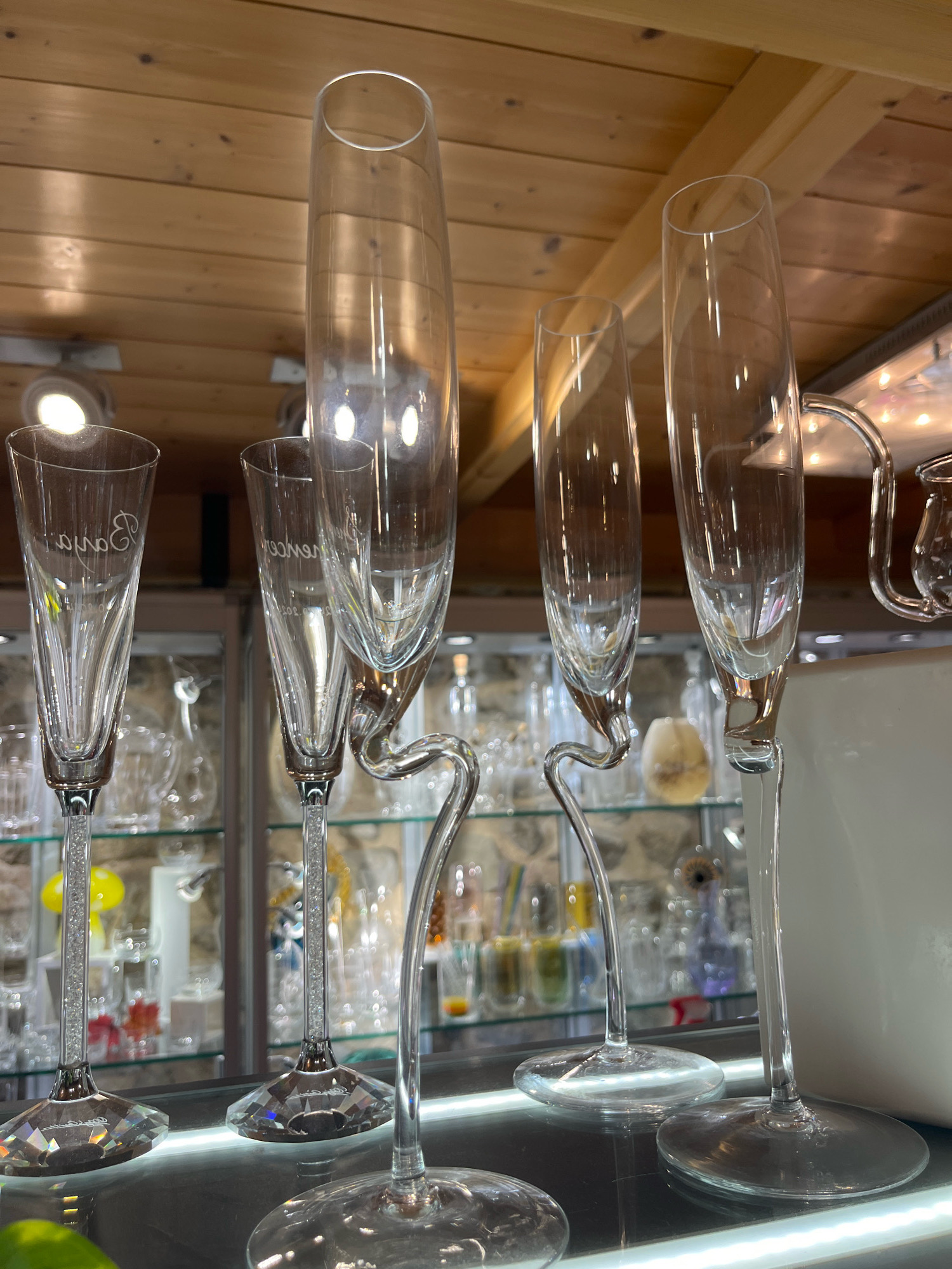 This image captures a close-up view of several champagne flutes displayed on a shelf within a shop. The flutes are made of clear glass and vary in design. Some have slender, elongated stems, while others feature more elaborate, curved stems that connect to the base. The bases of some of the flutes are decorative, encrusted with what appears to be small crystals, and faceted to refract light. The flutes are positioned in a line, with some overlapping each other. In the background, shelves lined with other glassware and decorative items are visible. The lighting suggests an indoor setting, possibly a retail environment. The ceiling above is made of wooden planks. The context of Yvoire, France, suggests this is likely a souvenir or gift shop in a tourist area, given the town's historical and picturesque reputation.