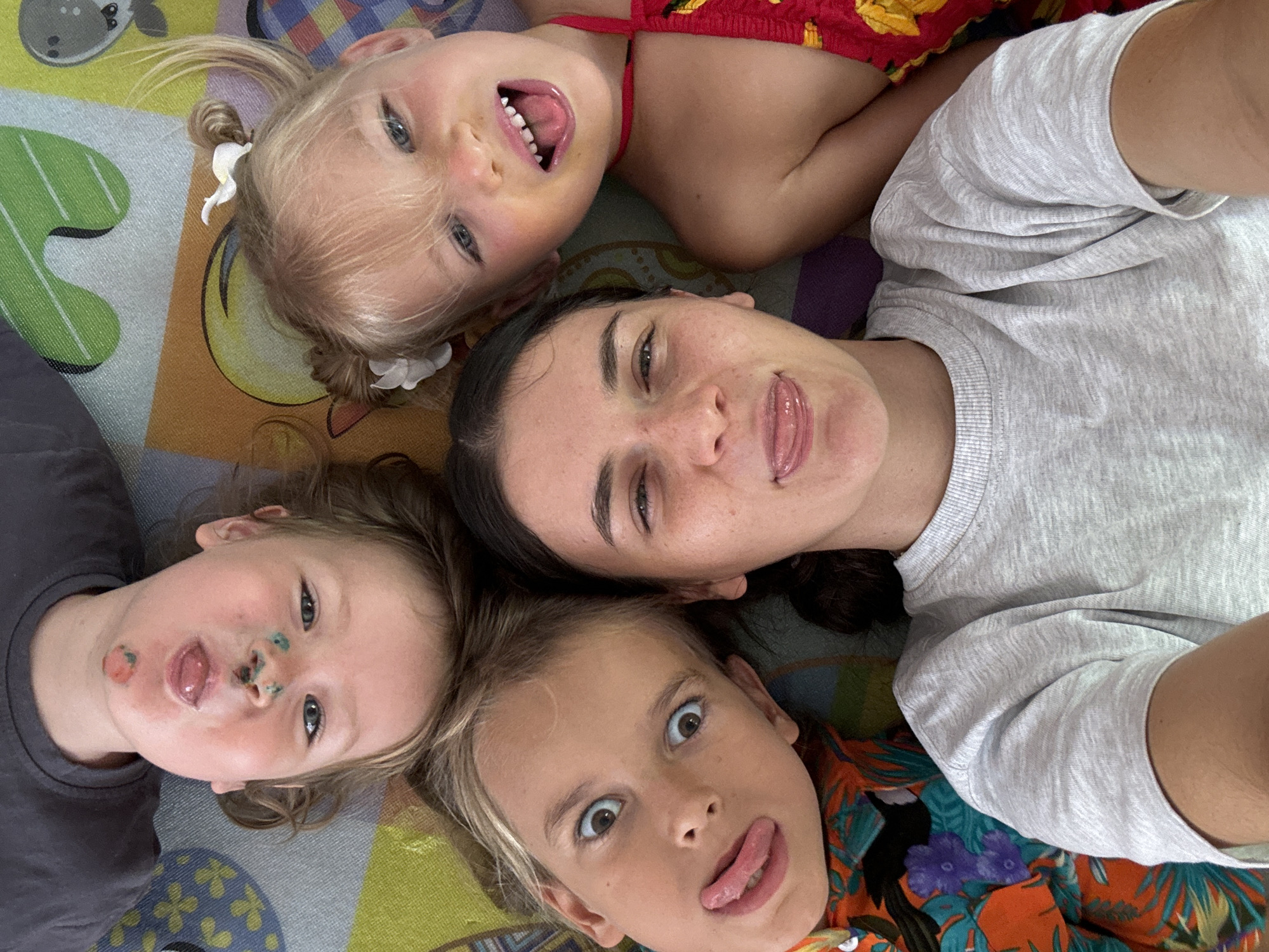 Four individuals are captured in a close-up selfie, likely taken indoors and situated on a patterned carpet or play mat. An adult female, wearing a grey t-shirt, is positioned centrally and appears to be taking the photo. She sticks out her tongue, mimicking the actions of some of the children.
The children, three in total, are positioned around the adult. At the top, a blonde-haired child in a red and yellow top is grinning, displaying their teeth with tongue sticking out. Another blonde-haired child at the bottom right also sticks out their tongue, expressing wide eyes and what looks like surprise. The final child at the bottom left also has their tongue out, but their face is marked with what appears to be remnants of food or paint. One of the children has white hair-clips.
The location of Mui Ne, Vietnam, is not identifiable within the visual information of the image.