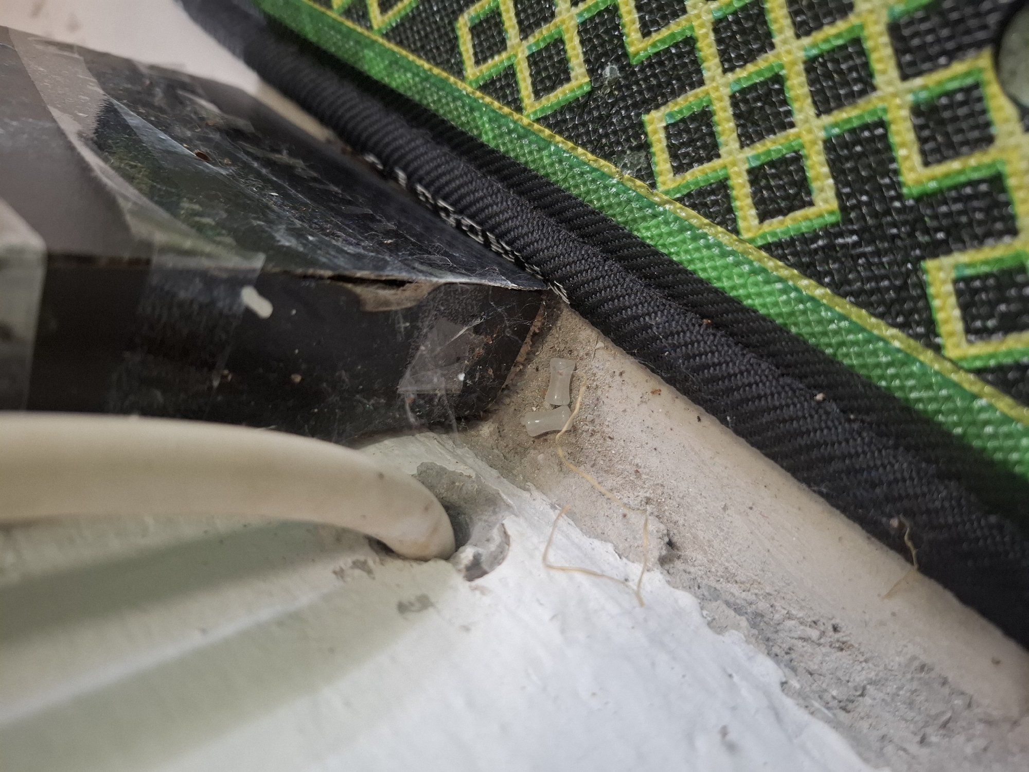 The image shows a close-up view of a wall and floor junction, with a cable entering the wall. A black electrical conduit or box is visible to the right, with some cobwebs attached. A section of a mat with a green and yellow geometric pattern is also in the frame. The scene appears to be indoors, possibly in a domestic or commercial setting. The lighting suggests an indoor environment. The overall impression is one of a mundane, everyday scene, with no particular event or action taking place. There is no information in the image to suggest the location in the city of Meureudu, Indonesia.