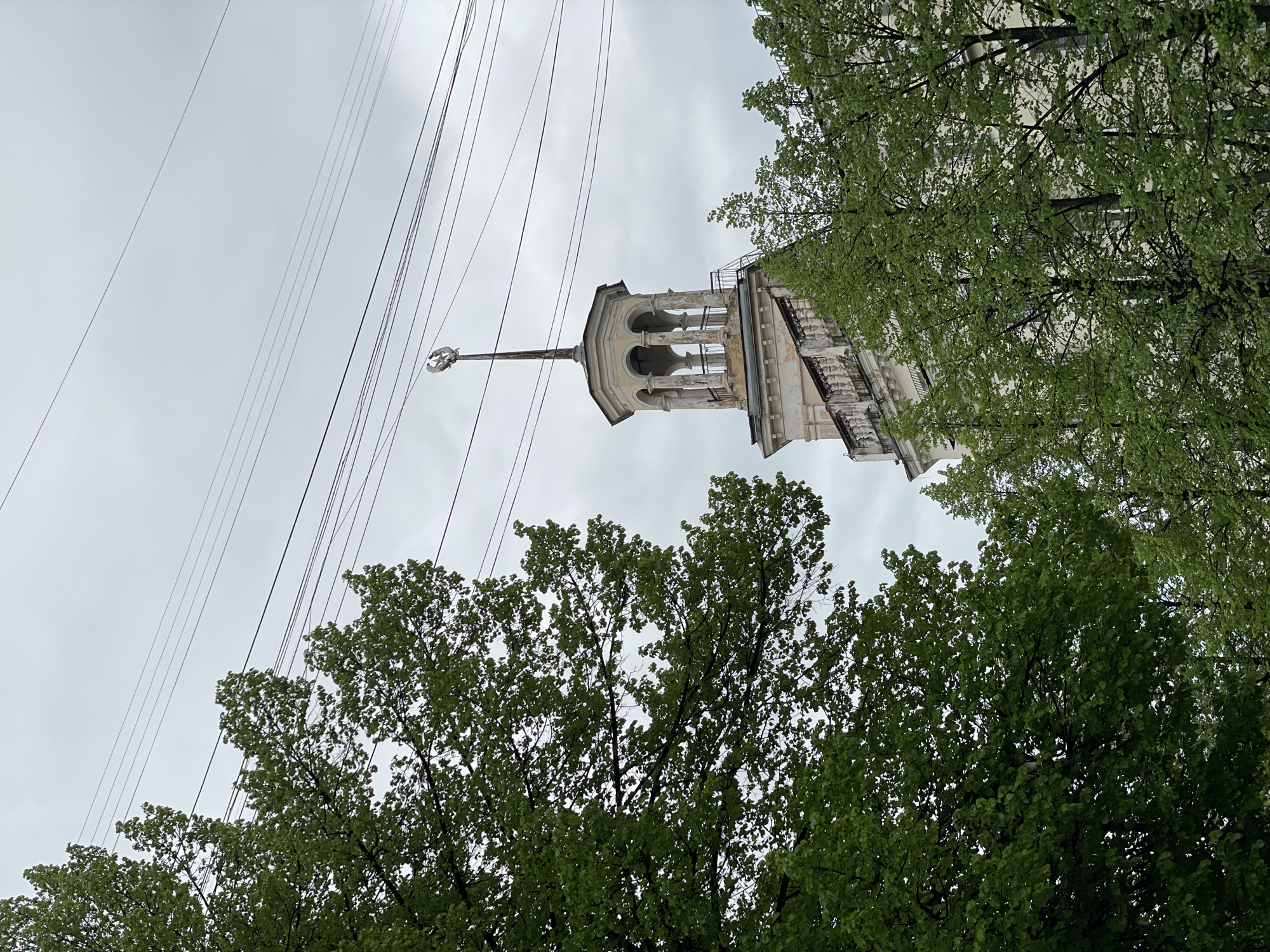 A low-angle view of a tall, multi-tiered architectural structure, likely a building tower, is partially obscured by trees. The tower is light in color with visible columns and a spire topped with a weather vane or decorative element. Power lines intersect the sky in the upper left quadrant of the image. The sky is overcast, and the trees have dense green foliage. The style and architectural details suggest the location is potentially Zheleznogorsk, Russia. The adjacent building to the tower has a visible staircase.