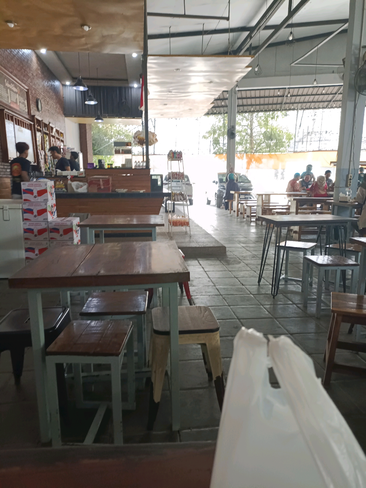 This is a photograph of a restaurant interior in Made, Indonesia, capturing a bustling scene. In the foreground, a white plastic bag obscures the lower portion of the image. Several wooden tables and chairs are visible, some with people seated at them. In the background, a counter area appears to be serving food and drinks, with staff members attending to customers. The restaurant has a rustic yet modern aesthetic, with exposed brick walls, wooden accents, and industrial-style lighting fixtures. The overall atmosphere suggests a lively and popular dining establishment.