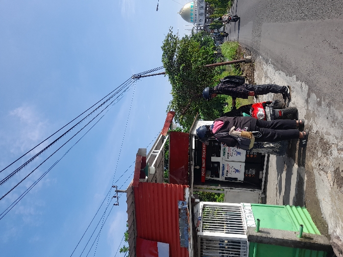 Two people on a motorcycle stand near a small roadside kiosk. The kiosk, with a red roof, displays the words "POM MINI DIGITAL." Overhead, power lines stretch across a clear blue sky. In the background, a building with a distinctive dome stands amidst lush greenery. The road curves into the distance, and a few other vehicles can be seen further along. The kiosk seems to be providing some service or product, possibly related to the motorcycle.