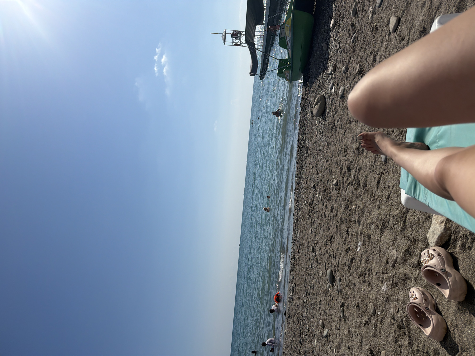 The image depicts a sunny day at a pebble beach in Sochi, Russia. In the foreground, a person's legs are visible, resting on a beach chair. Two pairs of light-pink clogs, embellished with pearls and starfish charms, are placed on the sand nearby. The beach itself is covered in dark pebbles and stones, leading to the clear blue water of the sea.
In the sea, several people are enjoying themselves. Some are wading near the shore, while others are swimming further out. One individual is wearing an orange life vest. Offshore, a green pedal boat with a canopy is docked, and further in the distance, a lifeguard tower is visible, along with other boats and a distant shoreline. The sky is a clear, vibrant blue, with a few wispy clouds and the sun shining brightly, casting a slight glare in the upper right corner. The overall scene conveys a relaxed and leisurely atmosphere typical of a summer day at the beach.