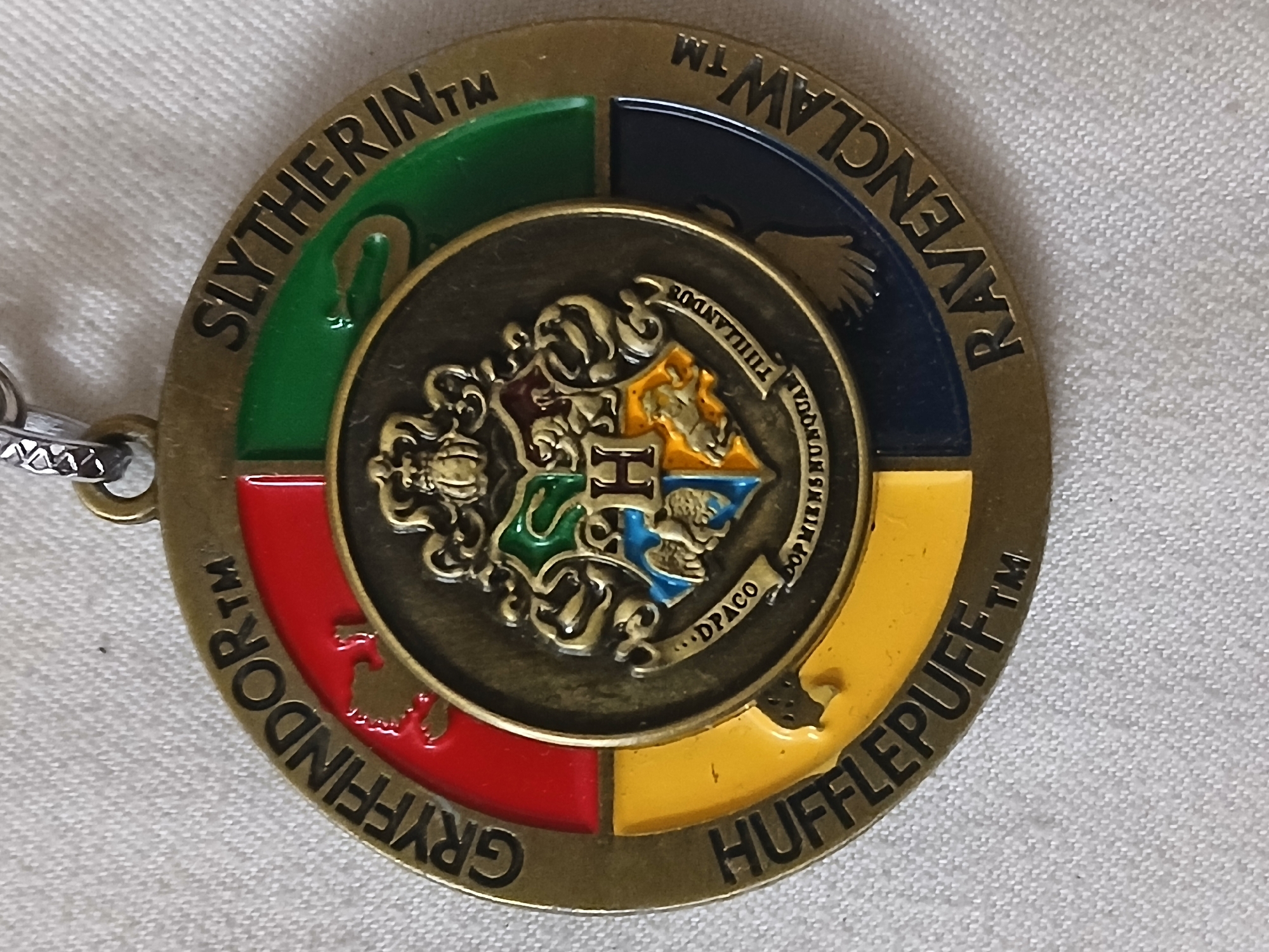 The image depicts a round, metallic keychain featuring the Hogwarts school crest from the Harry Potter series. The keychain is divided into four quadrants, each representing one of the Hogwarts houses: Slytherin (green), Ravenclaw (navy blue), Hufflepuff (yellow), and Gryffindor (red). The house names are printed in black around the outer edge of each quadrant. In the center of the keychain is a smaller, raised circle depicting the Hogwarts school crest in detail, featuring the animals representing each house and the letter "H". The keychain is attached to a chain on the left side. The background is a white, textured surface. There is no information that confirms the location is Bengaluru, India.