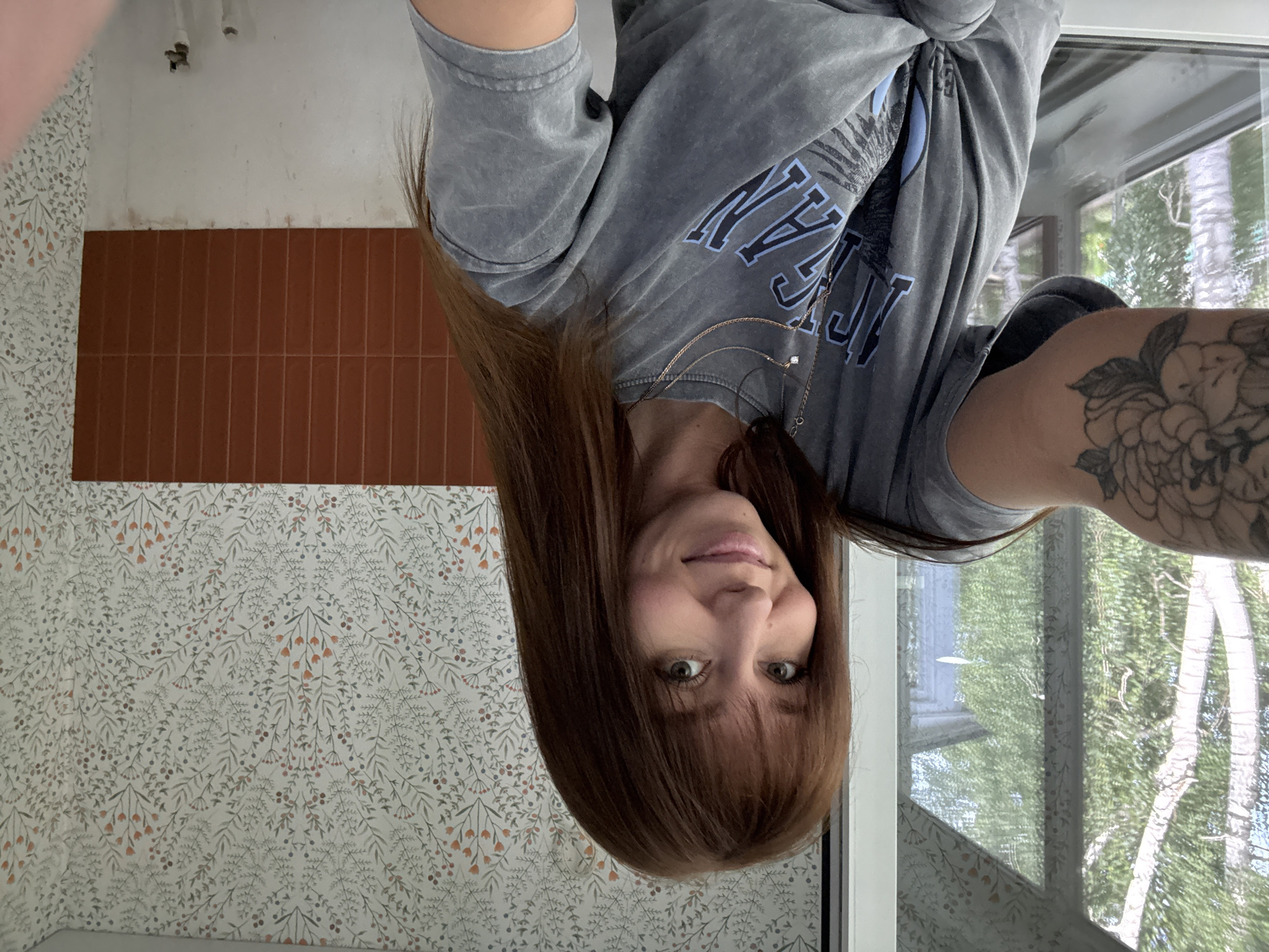 This is a portrait-oriented, vertical, selfie-style photograph taken by a person with long, brown hair and bangs. They are wearing a grey, long-sleeved t-shirt with blue lettering on the chest and a subtle floral tattoo on their upper left arm. The person's face is turned towards the camera, with their head tilted slightly. They have green eyes and a gentle smile.
The background shows a wall with floral wallpaper featuring a pattern of light green leaves and small orange and grey berries. Above the wallpaper, there is a section of terracotta-colored rectangular tiles. To the right of the person, a large window reveals an outdoor scene with lush green trees, suggesting a daytime setting. The lighting in the image is bright, indicating daylight, and there are no obvious signs of specific weather conditions. The overall impression is casual and relaxed. The person appears to be indoors, possibly at home, given the domestic setting.