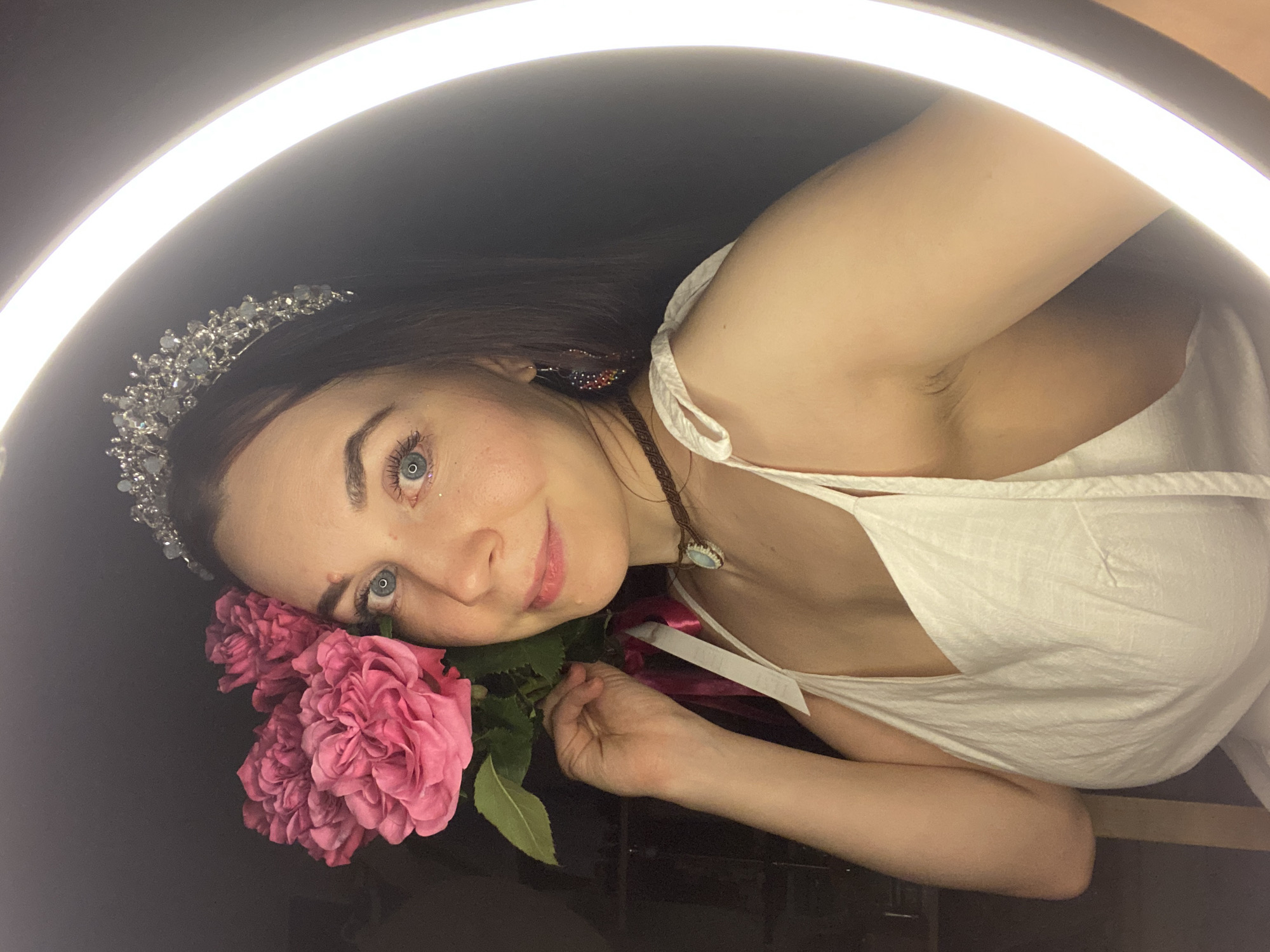 A woman with long brown hair is posing with pink flowers under a circular light. She is wearing a white dress with a tie shoulder and a tiara. She also has a necklace, and her eyes are blue. The background is dark. There is no indication of the city Chaikovsky, Russia.