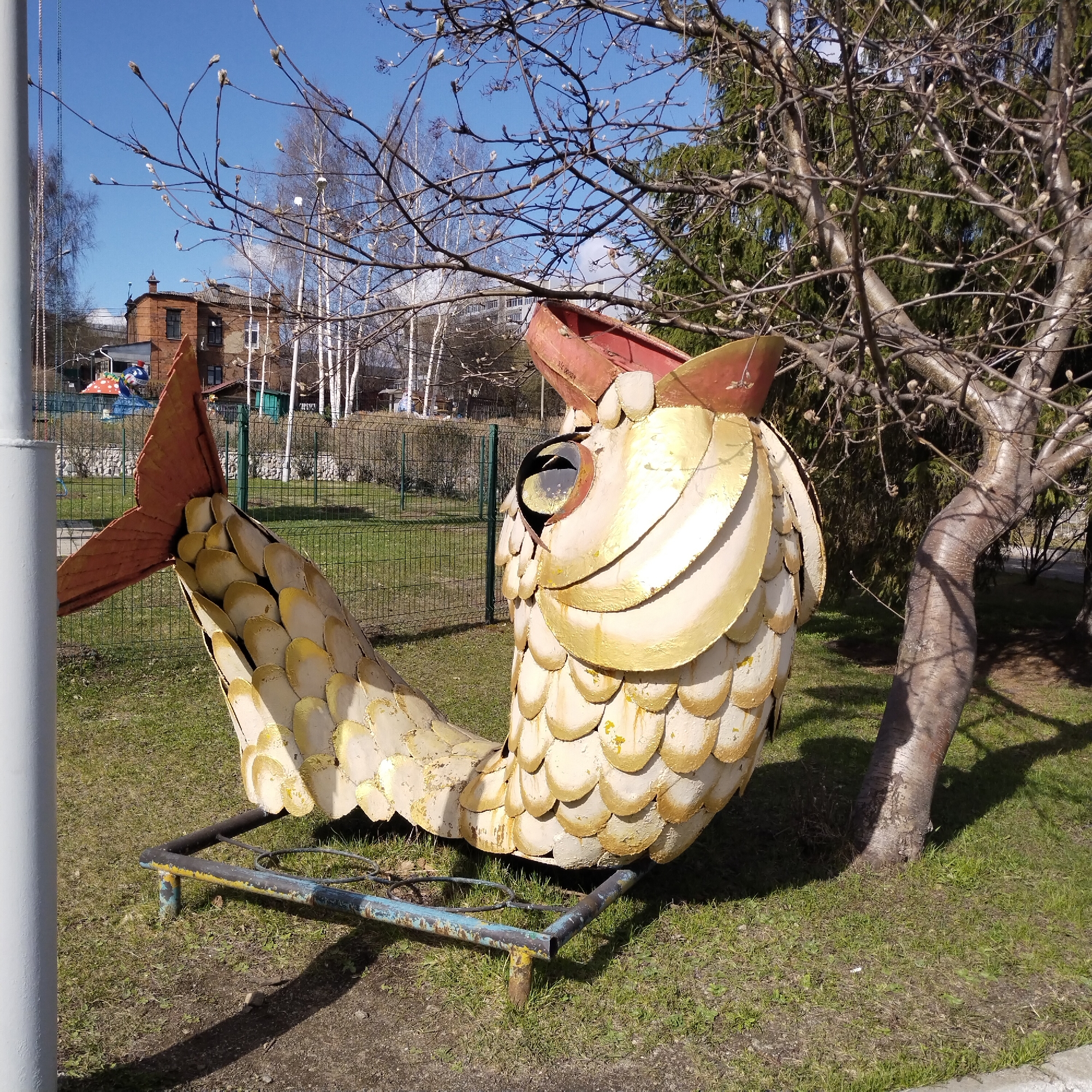 A full shot captures a large, weathered, metal fish sculpture, mounted on a rusted metal frame on a grassy patch. The fish sculpture is predominantly off-white with yellow accents, styled with overlapping scale-like plates. It has a large, circular eye socket and a reddish-brown tail. The sculpture appears aged with noticeable wear and tear.
To the right of the sculpture is a small tree with bare branches, some with budding leaves. The tree trunk is light gray and stands against a backdrop of green foliage.
In the distance, behind a metal fence, stands a red brick building with white trim. There are also several birch trees with white bark visible in the background, under a clear blue sky.
The location is inferred as Nizhny Tagil, Russia.