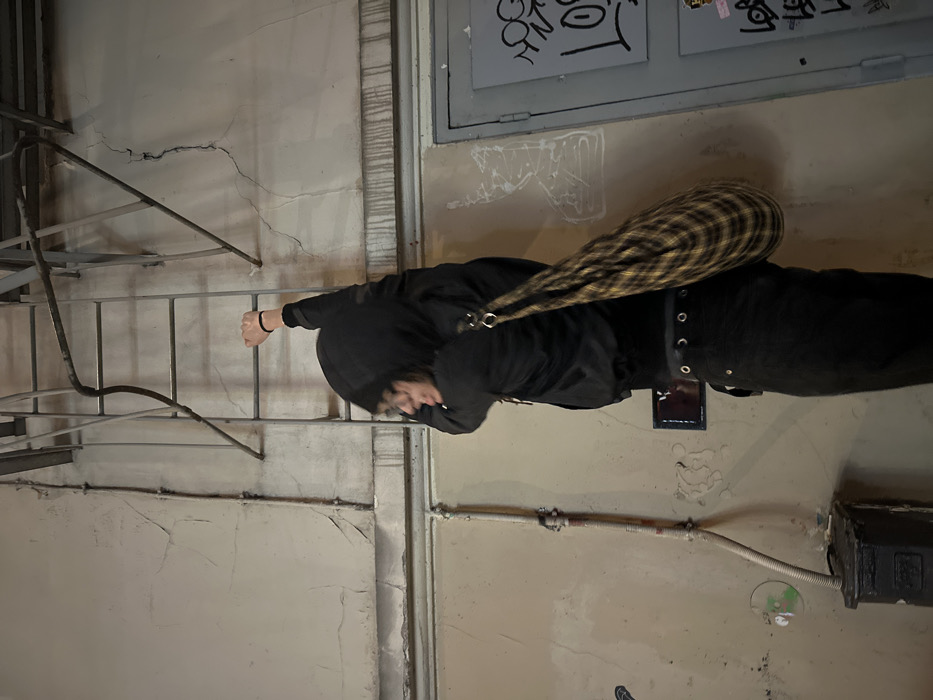 A person in a black hooded jacket and black jeans stands near a metal railing. The person carries a yellow and black plaid bag over their shoulder. The setting is outdoors, against a wall with pipes and electrical boxes. Graffiti marks a section of the wall above. The overall scene is dimly lit, suggesting a nighttime setting.