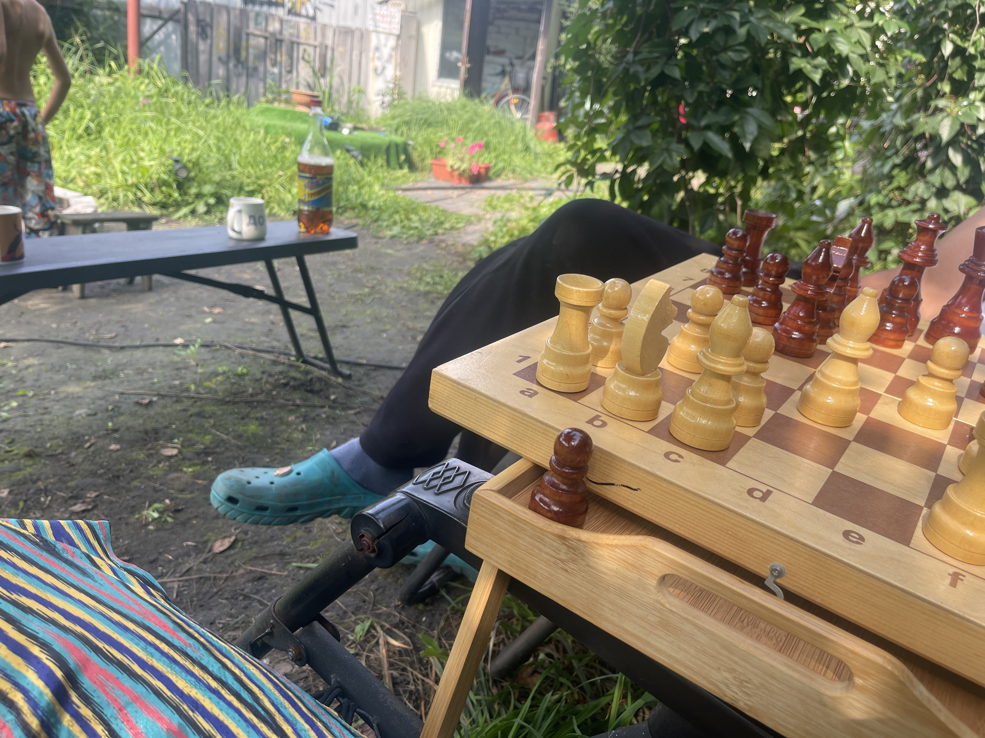 In Barnaul, Russia, on a sunny day, a person with vibrant patterned shorts stands in the background, their back to the camera. In the foreground, a chess game is in progress on a wooden board. The pieces are a mix of light and dark wood, with some pieces already captured and resting in a small compartment of the board. The board is set up on a portable table, which appears to be set up outdoors in a yard or garden area. Lush green vegetation surrounds the scene, with a bottle of amber liquid and a white mug visible on a nearby table. The ground is a mix of dirt and grass, suggesting a casual, outdoor setting. A teal Croc shoe is visible on the foot of the person playing chess, which is partially obscured by a blanket with a colorful striped pattern. The overall impression is one of a relaxed outdoor activity, perhaps a friendly game of chess on a warm day in Barnaul.