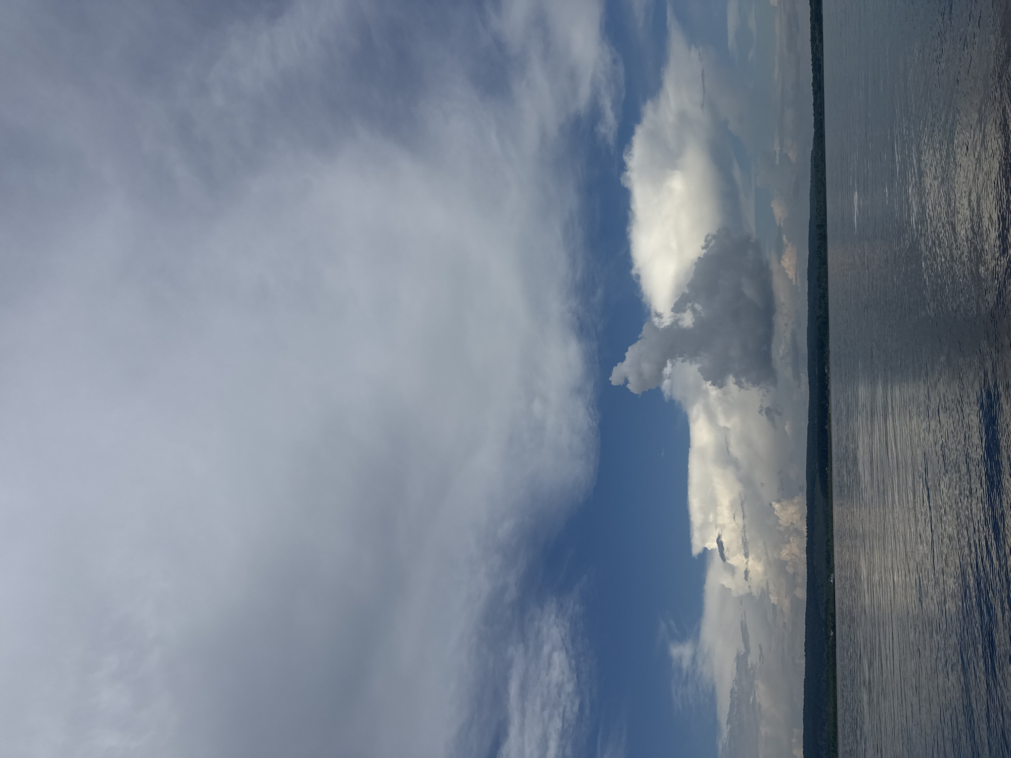 The image shows a wide, calm body of water in the foreground, with gentle ripples on its surface reflecting the sky. A distant, dark green treeline stretches across the horizon, with a few subtle indications of structures nestled within it. Above the treeline, a dramatic sky dominates the scene. Large, fluffy white clouds are interspersed with patches of deep blue sky, and wispy, grey clouds create a layered effect. The overall impression is one of a serene, natural landscape under a dynamic, cloud-filled sky. The location is described as Unknown, Unknown.