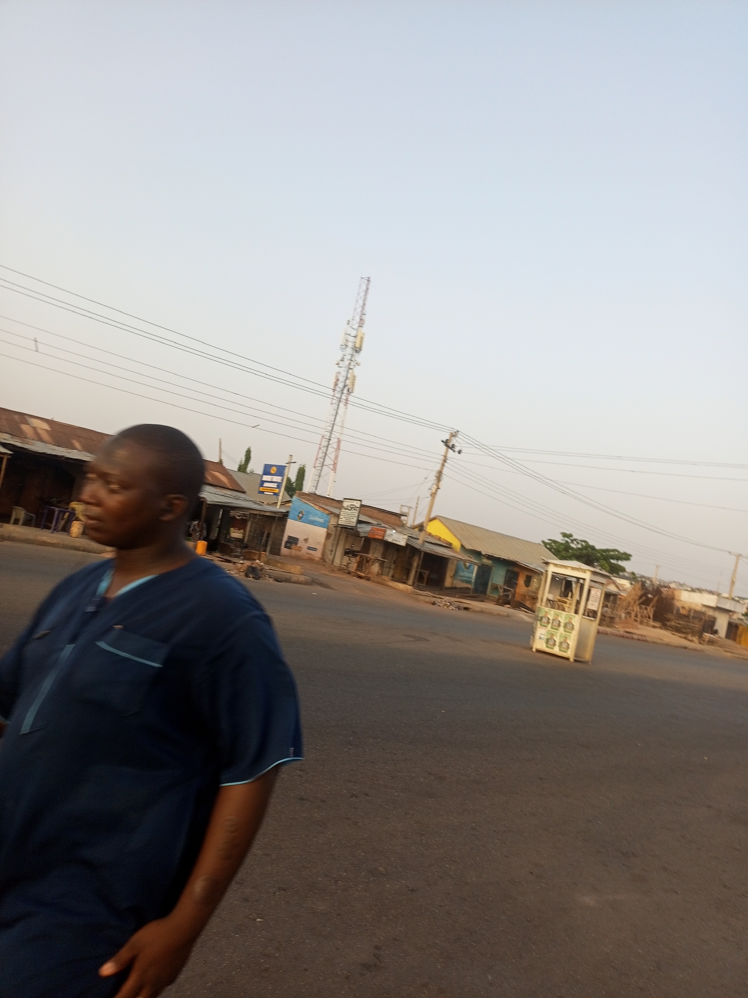 The image shows a street scene. A man is walking on the road, facing towards the left of the frame. Behind him, there are buildings and structures along the side of the road. A cell tower stands tall in the background. Utility poles with wires run above the buildings. A small white booth is visible on the right side of the road. The sky appears clear with a gradient of light color. The location is the city of Unknown, Unknown.