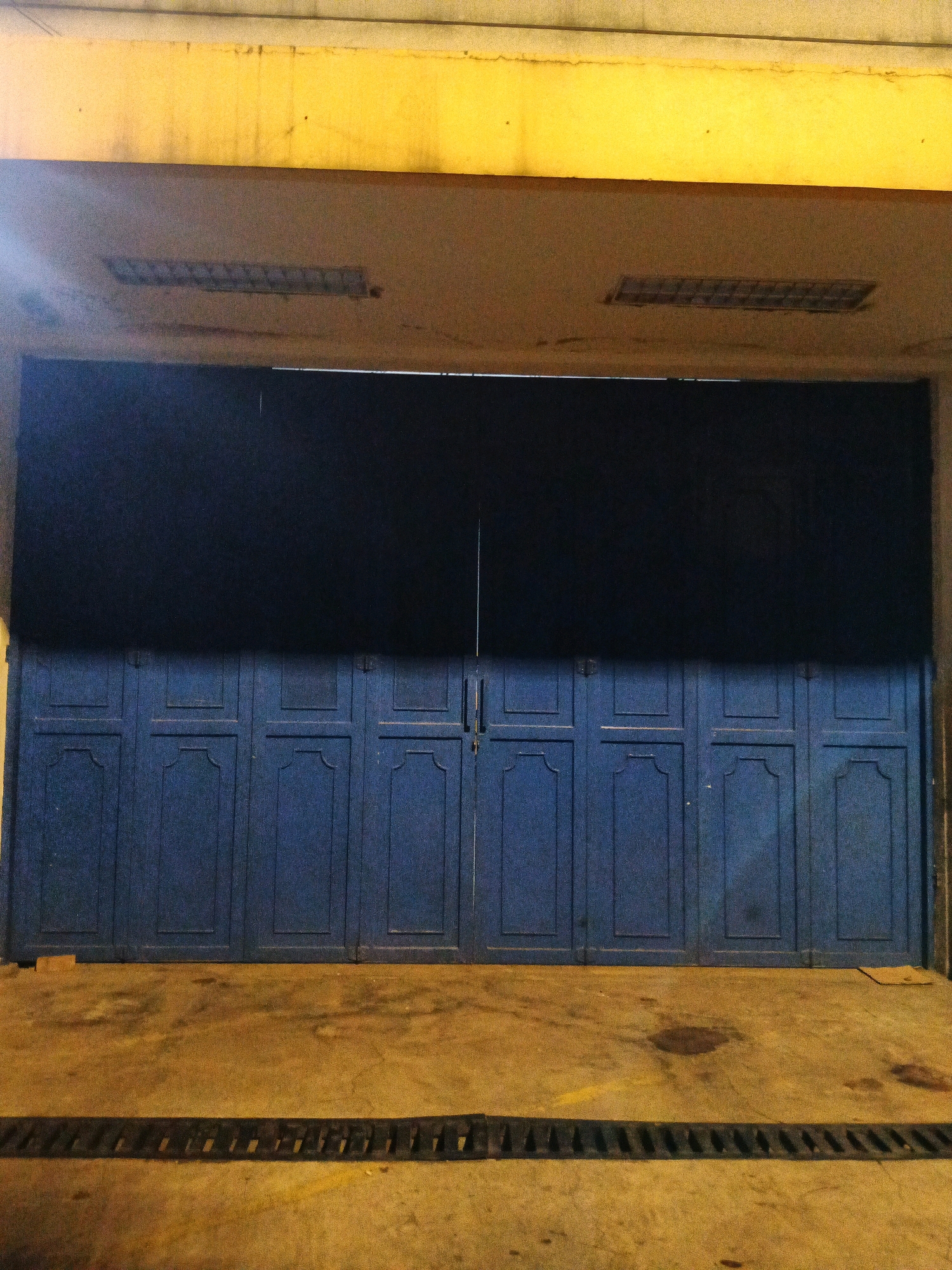 The image shows a large, dark blue, corrugated metal gate with six panels, blocking the entrance to a building. The gate has decorative molding on each panel. Above the gate, a yellow, textured awning is visible, with two rectangular fluorescent light fixtures mounted on its underside. The ground in front of the gate is a concrete surface with some stains and a black, grated drain cover running horizontally across it. The scene appears to be in an urban setting, possibly a commercial or industrial area, given the large gate and lighting fixtures. The overall lighting is dim and appears to be artificial, casting a yellow hue.