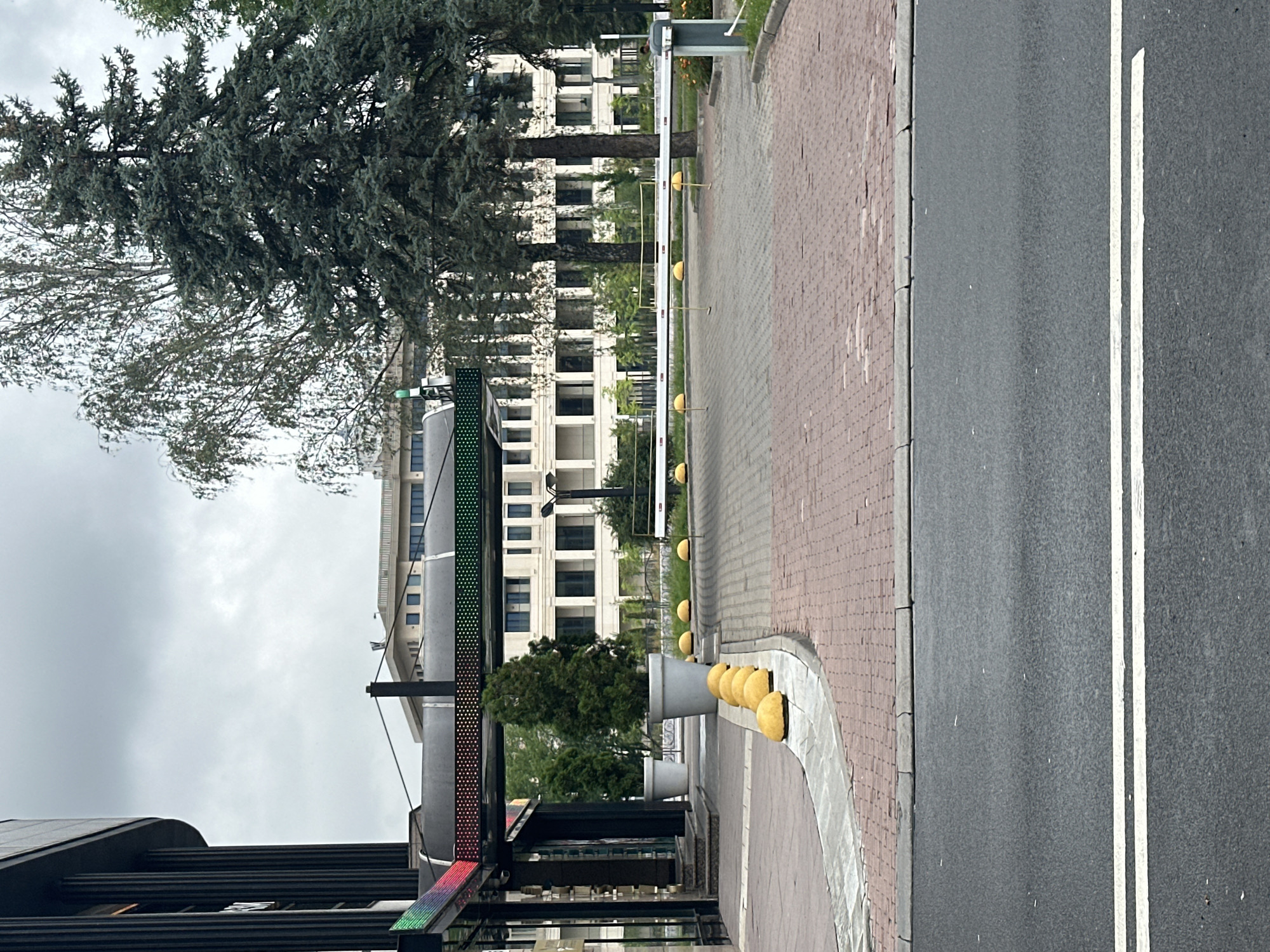 The image depicts an outdoor scene in what appears to be a modern urban environment, possibly in Minsk, Belarus, characterized by a mix of contemporary architecture and natural elements. In the foreground, a paved road with double white lines indicates a clear traffic lane. Adjacent to the road is a brick-patterned sidewalk or pedestrian area. This area is demarcated by a concrete curb and lined with several rounded, yellow bollards.
The midground reveals an entrance area to a building, possibly a commercial or office complex. A dark-colored modern building with vertical design elements is visible on the left. Above the entrance, a covered walkway or canopy is present, adorned with a strip of colorful LED lights displaying a gradient from red to green. Large, white planter pots containing evergreen trees are placed along the walkway.
Further back, a white barrier arm, commonly used for vehicle access control, is extended across a paved area. This area also features yellow bollards along its perimeter. Behind the barrier, a multi-story building with a light-colored facade and numerous windows is visible, suggesting it is part of a larger complex. Mature trees, including a tall evergreen and a deciduous tree with light-colored foliage, stand between the entrance and the background building, providing a natural contrast to the architecture. The sky is overcast, suggesting a cloudy day. No people are clearly visible in the image.