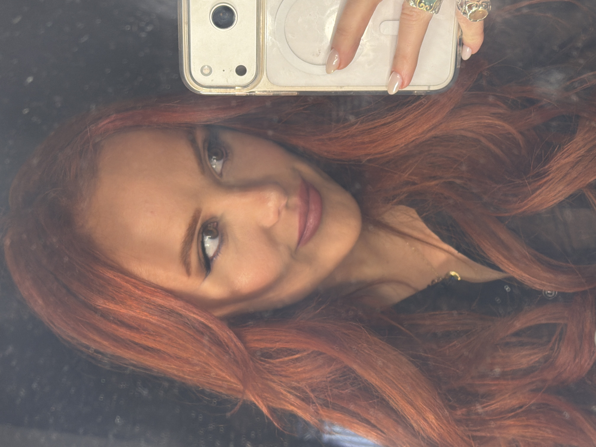 A selfie shot shows a woman with reddish-brown hair taking a picture in a reflective surface. She is holding a white smartphone with rings on her fingers. The background is blurred. The location is not identifiable as Paris, France.